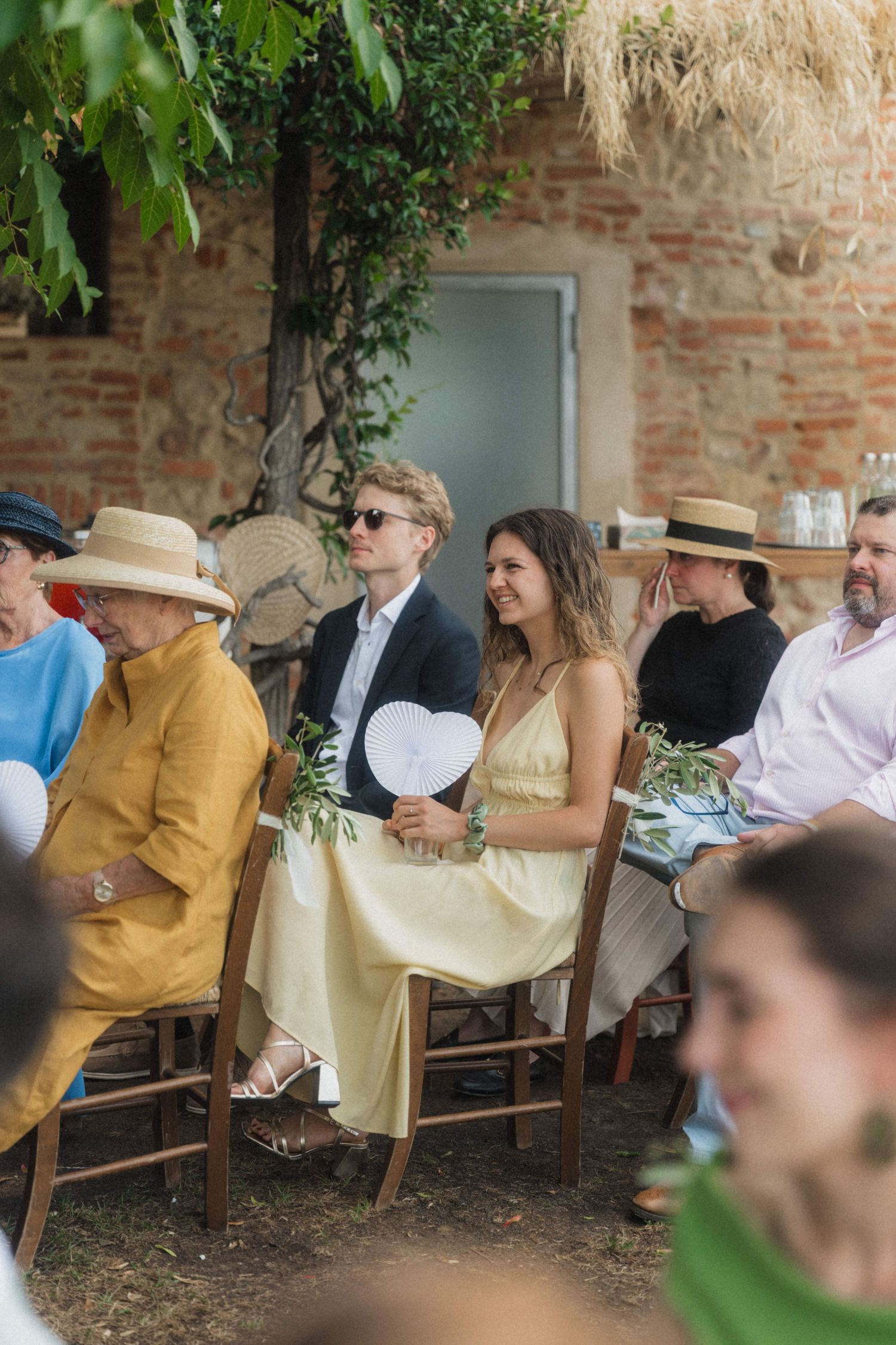 Wedding guests seated outdoors, woman in yellow dress holding heart-shaped fan at sunny ceremony.
