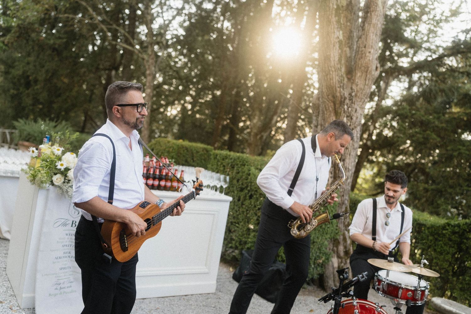 Live wedding band performing outdoors with guitarist, saxophonist, and drummer in golden sunlight.