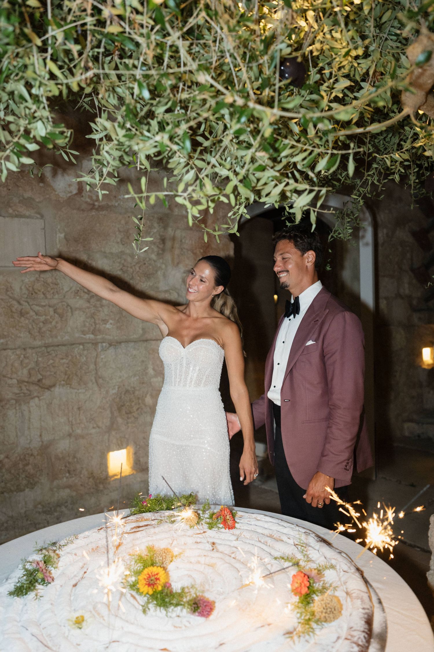 Smiling bride in a beaded white gown and groom in a burgundy blazer beside a floral wedding cake topped with sparklers under olive branches.