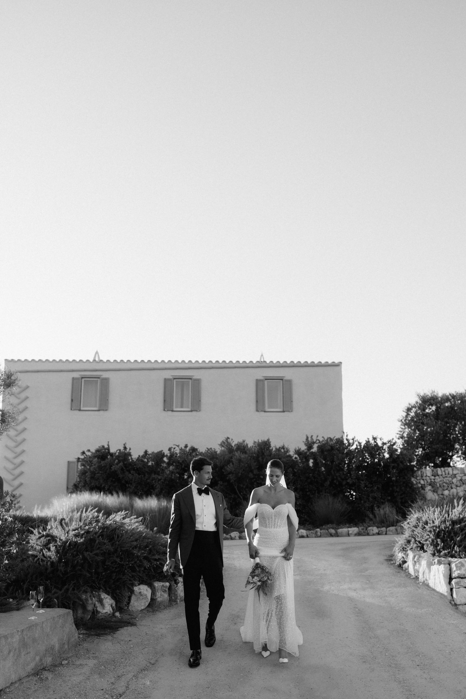 Black and white photo of the bride and groom walking hand in hand toward a Mediterranean stone house.