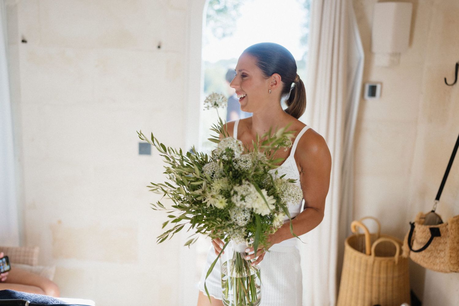 Smiling bride in a white dress holds a large bouquet of white and green wildflowers in a bright room with wicker baskets.