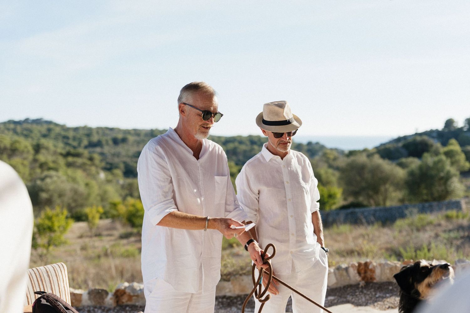 Two men in white linen outfits and sunglasses stroll with a dog on a leash outdoors, with green hills in the background.
