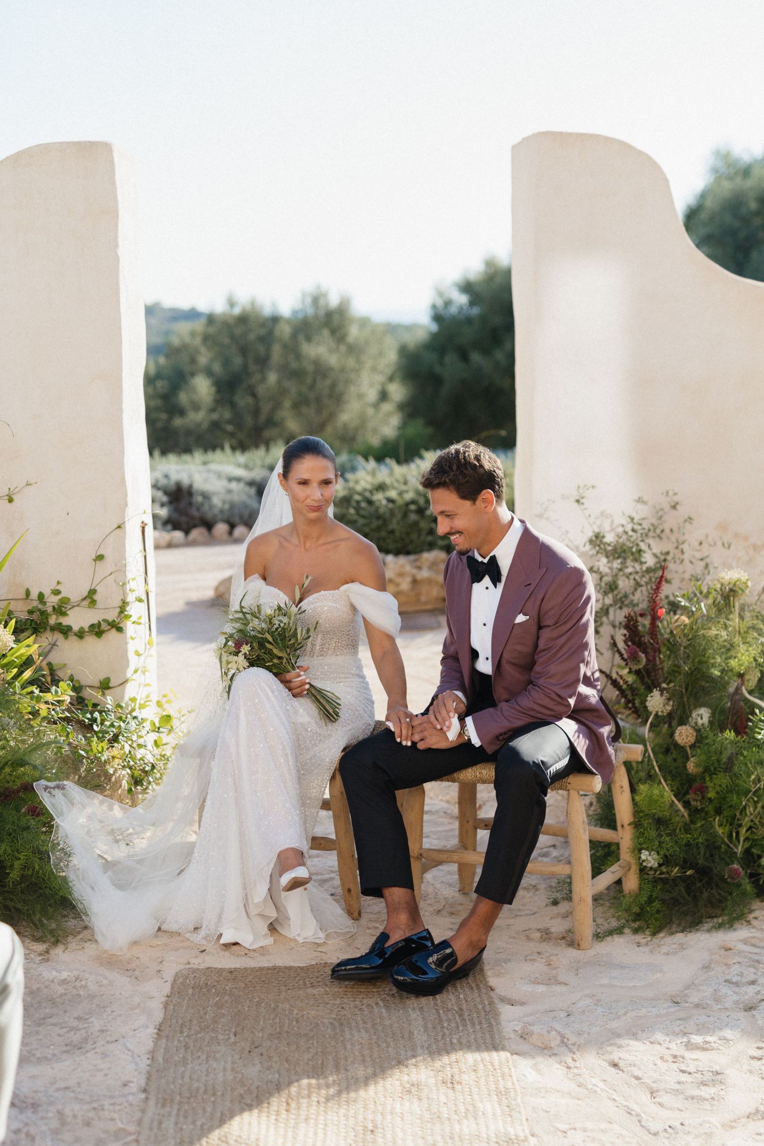 Bride in an off-shoulder lace gown and groom in a burgundy blazer hold hands on wooden stools at an outdoor ceremony.