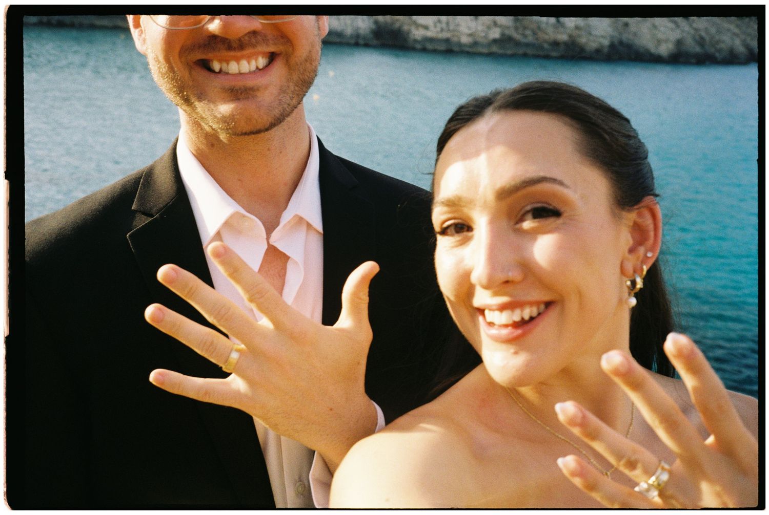 Smiling couple pose by the sea as the woman holds up her hand to show off a sparkling engagement ring, celebrating their recent engagement.