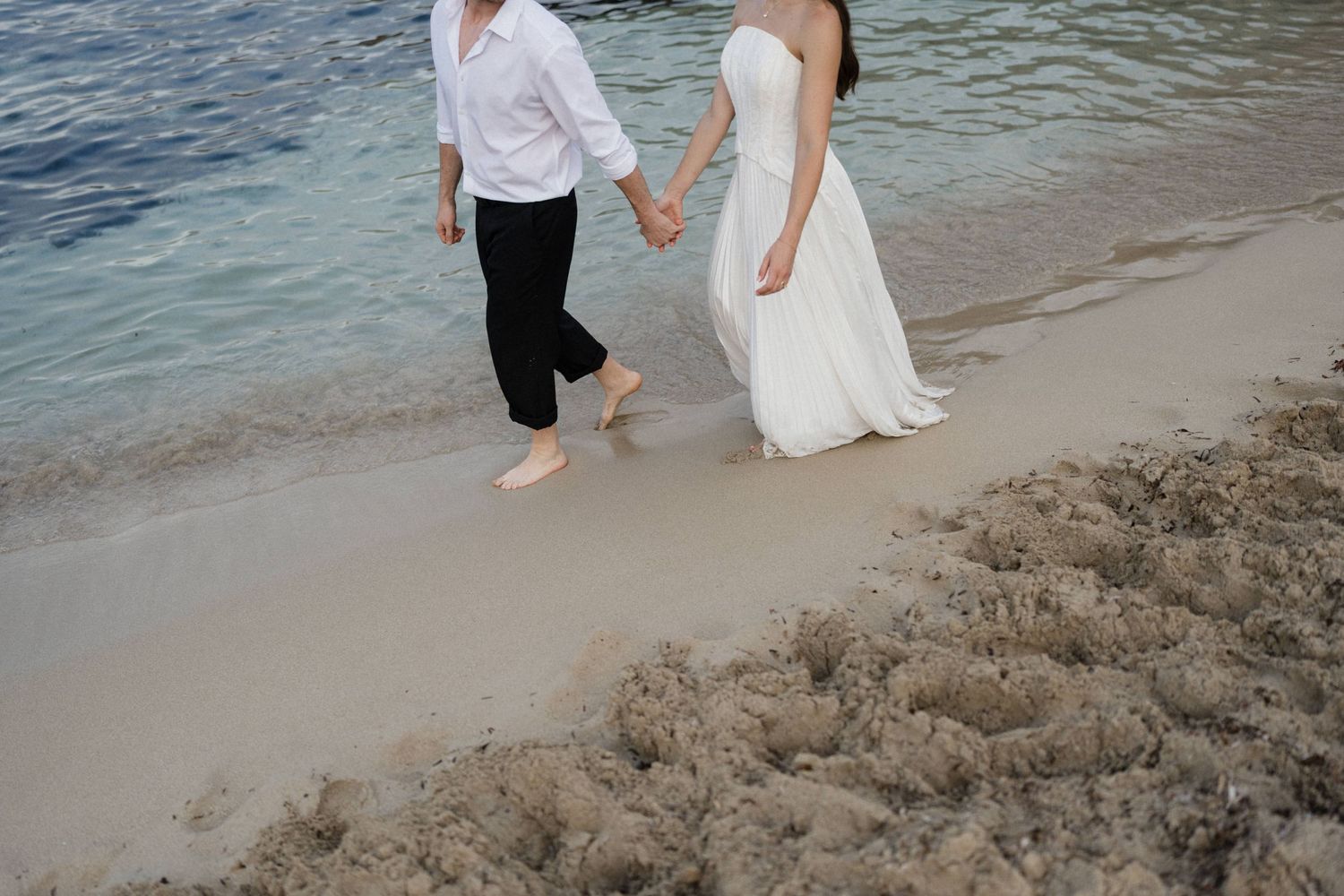 Barefoot couple in white dress and black pants hold hands while walking along a sandy tropical beach beside gentle turquoise waves.