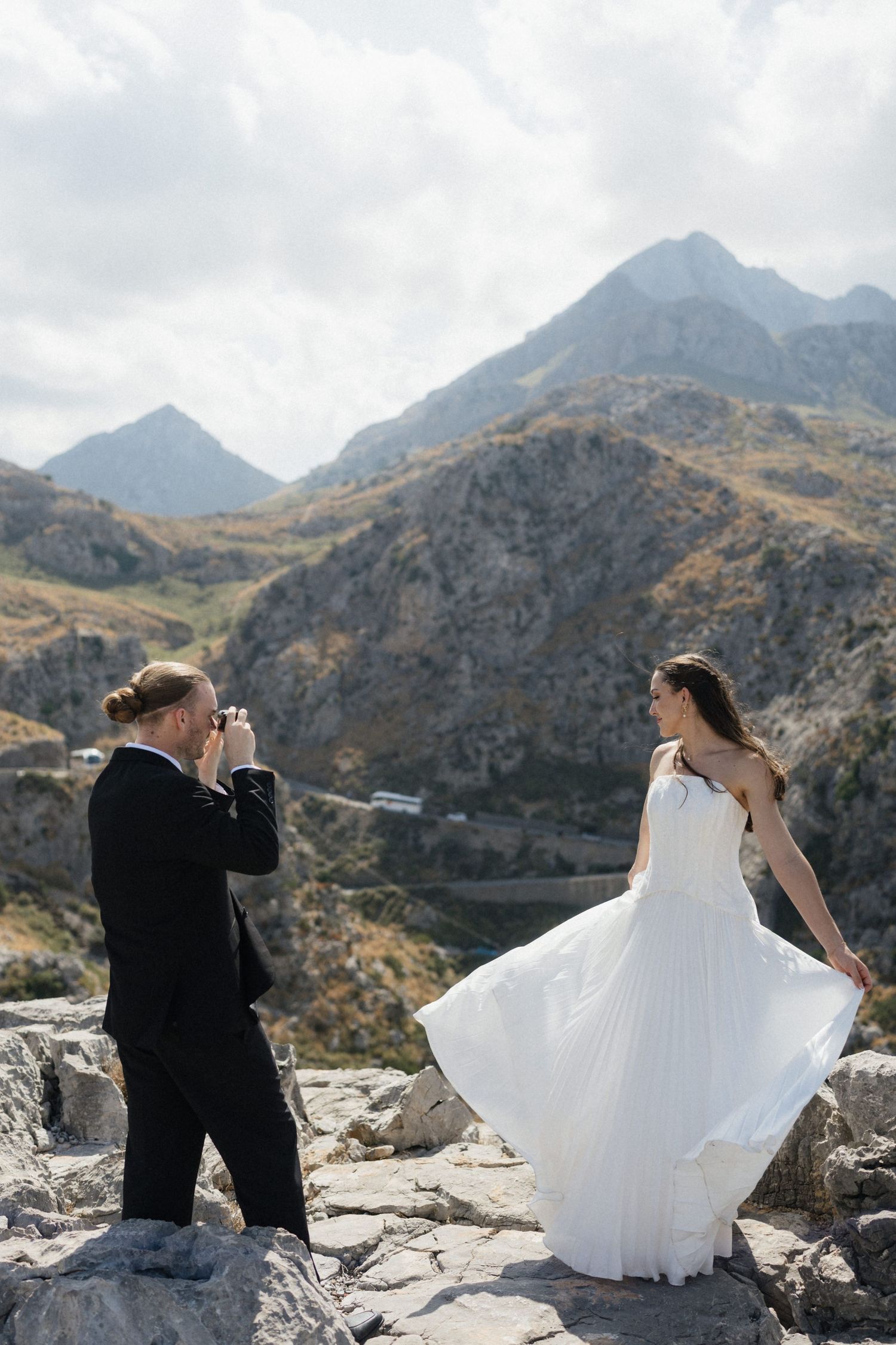 Groom in a black suit photographs a bride in a flowing white wedding dress on a rocky mountain overlook during a scenic destination wedding.