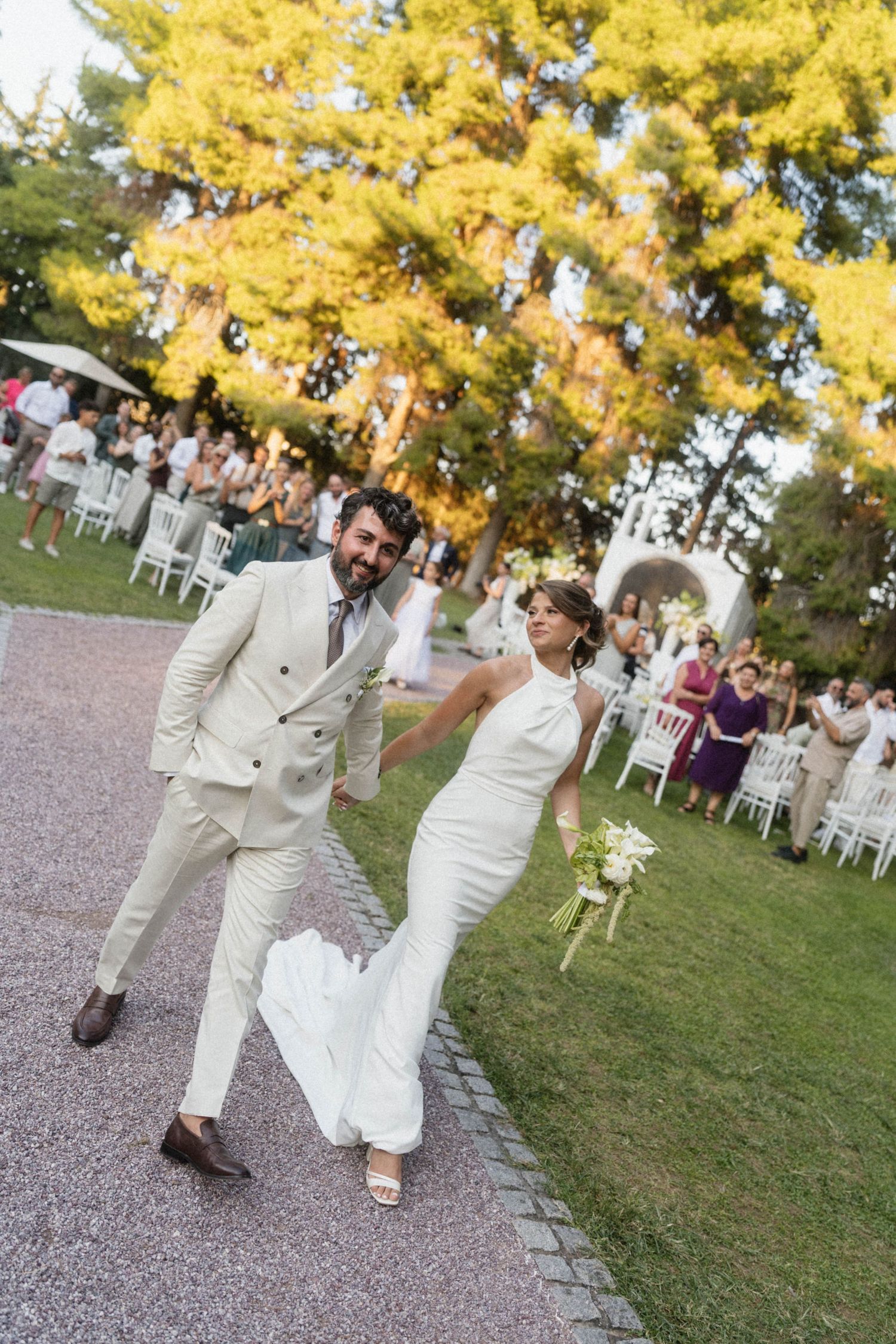 Joyful newlyweds walk down outdoor aisle, bride in white gown holding bouquet, groom in cream suit, guests seated on chairs.