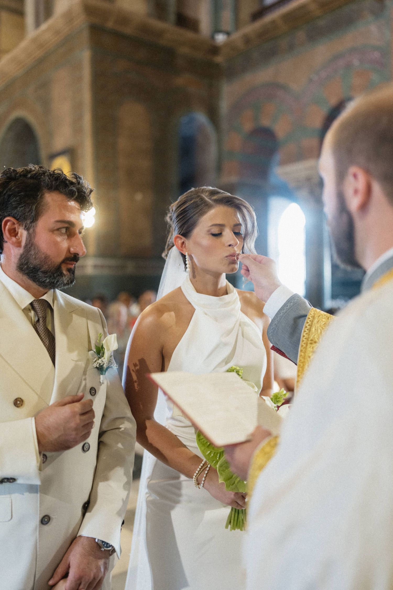 Bride and groom receive blessing from Orthodox priest during elegant church wedding ceremony.