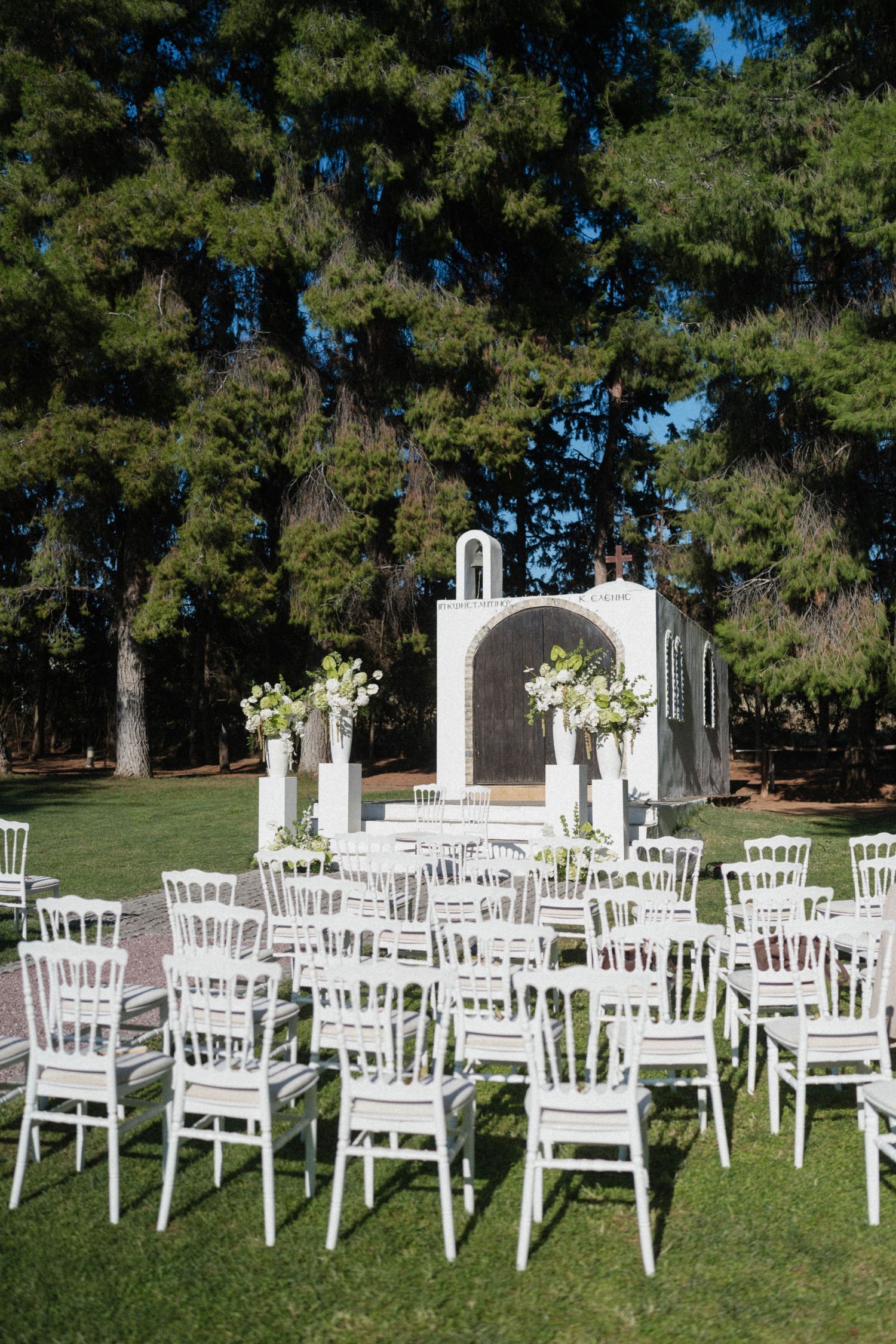 Elegant outdoor wedding ceremony setup with white chairs and floral arrangements on green lawn with pine trees.