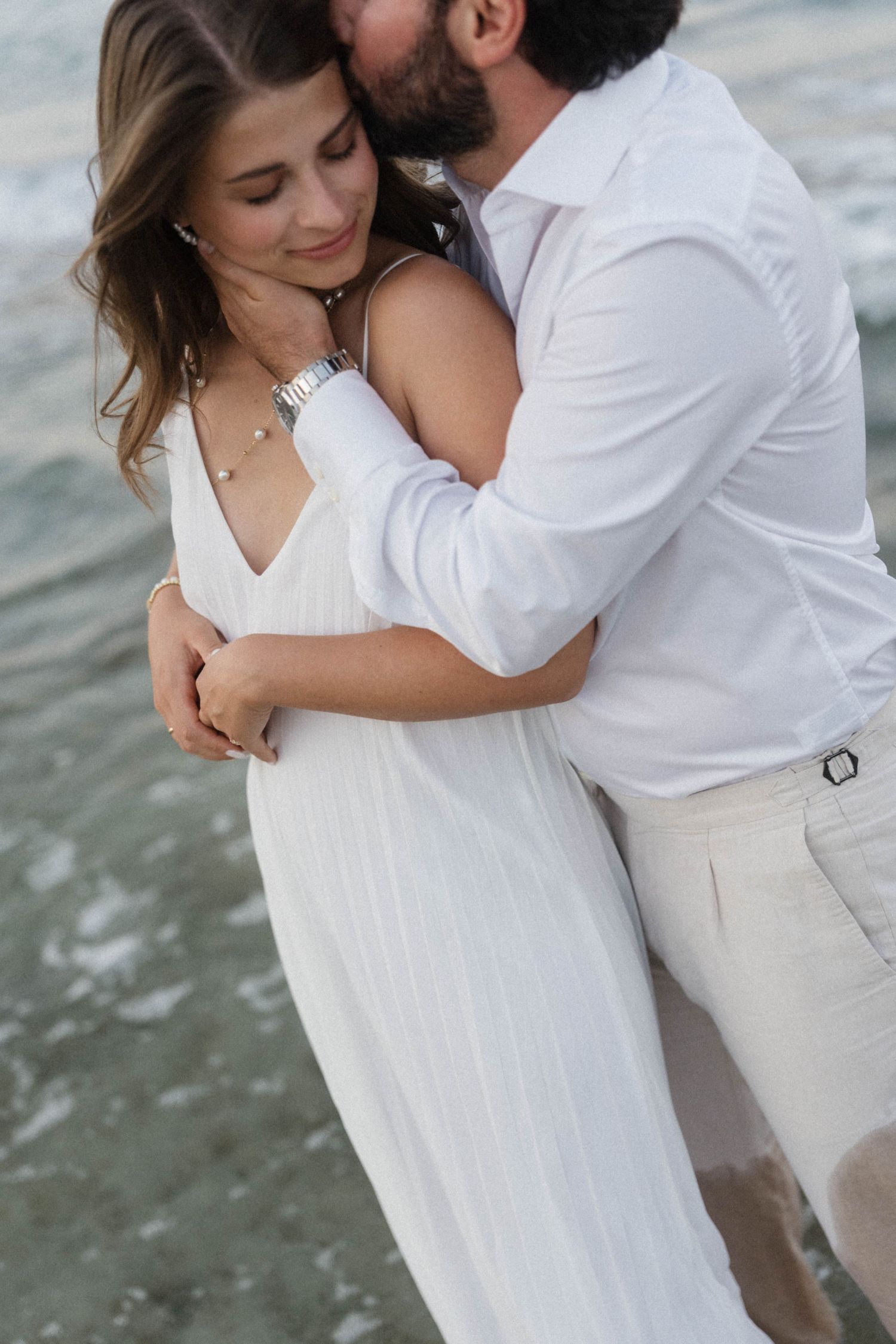Romantic couple embracing on beach shore, woman in white dress smiling as man kisses her cheek near ocean waves.