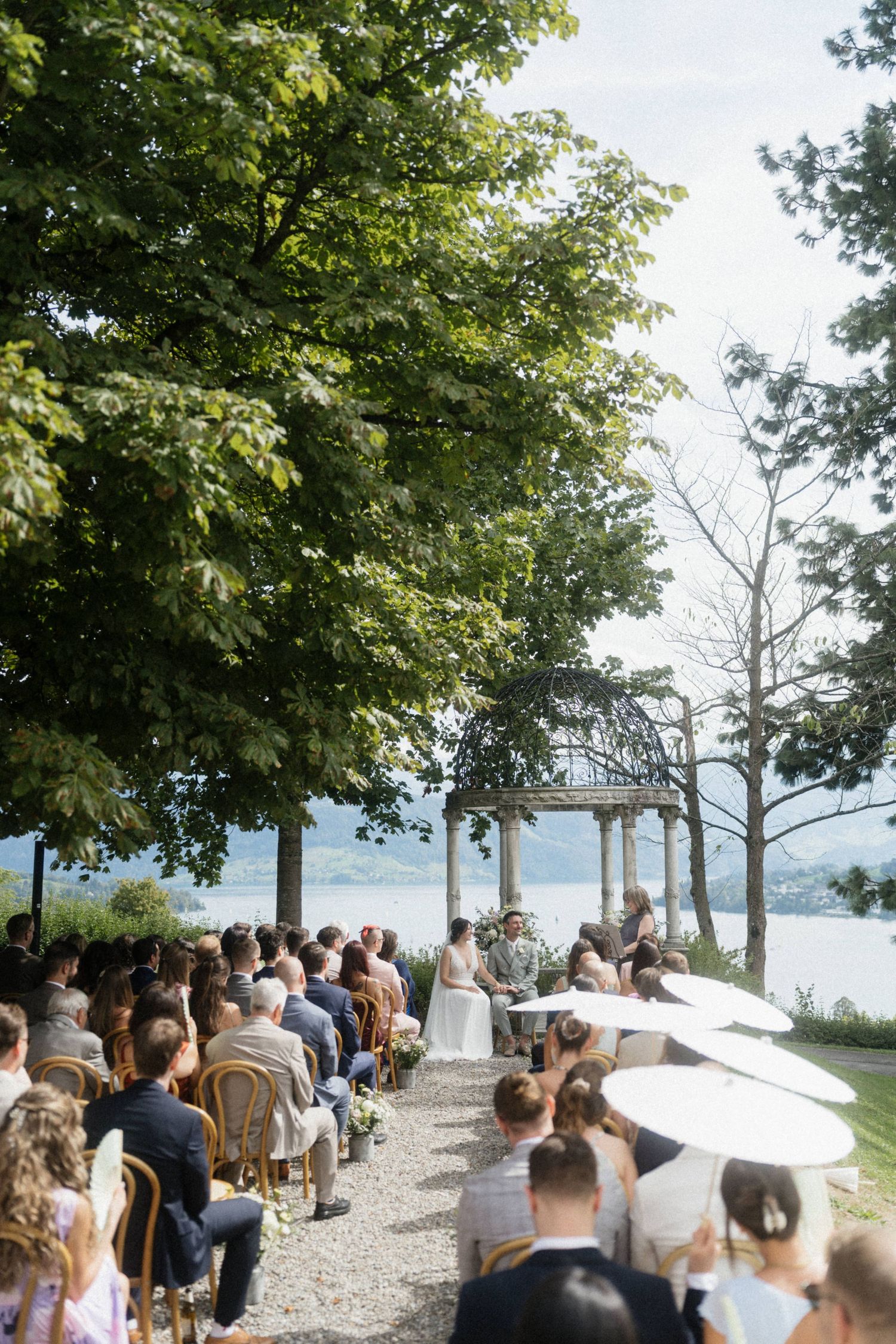 Outdoor lakeside wedding ceremony with guests seated on golden chairs beneath lush trees with scenic water views.