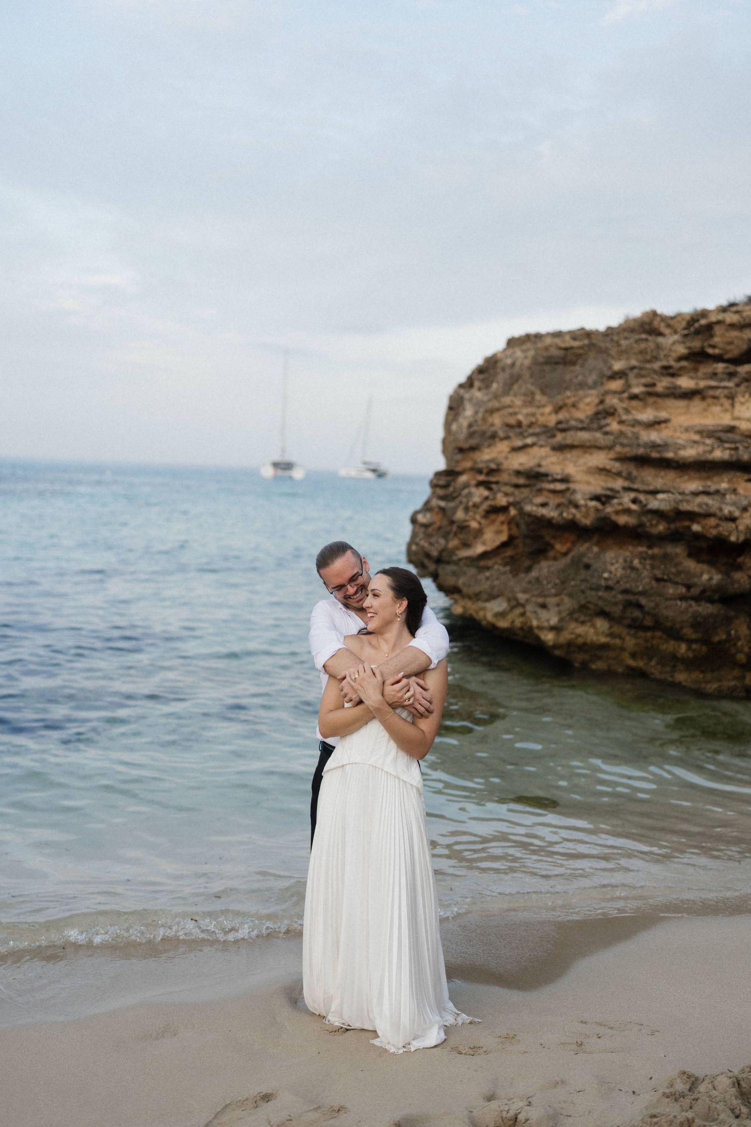 Couple embracing on a sandy beach at the water's edge, the bride in a flowing white gown, with rocky cliffs and sailboats behind.