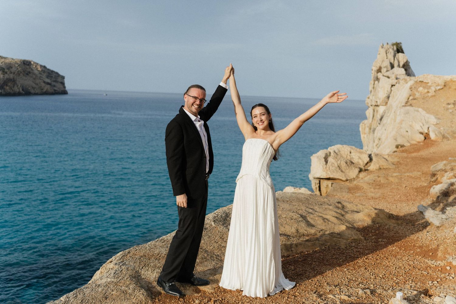 Bride in a white gown and groom in a dark suit raise joined hands in celebration on a rocky seaside cliff overlooking the blue sea.