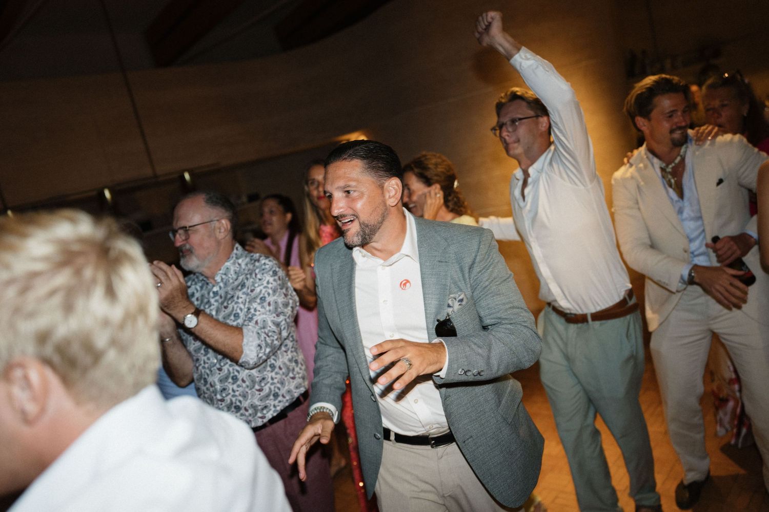 A smiling man in a gray blazer dances with guests, hands raised, at a lively wedding party.