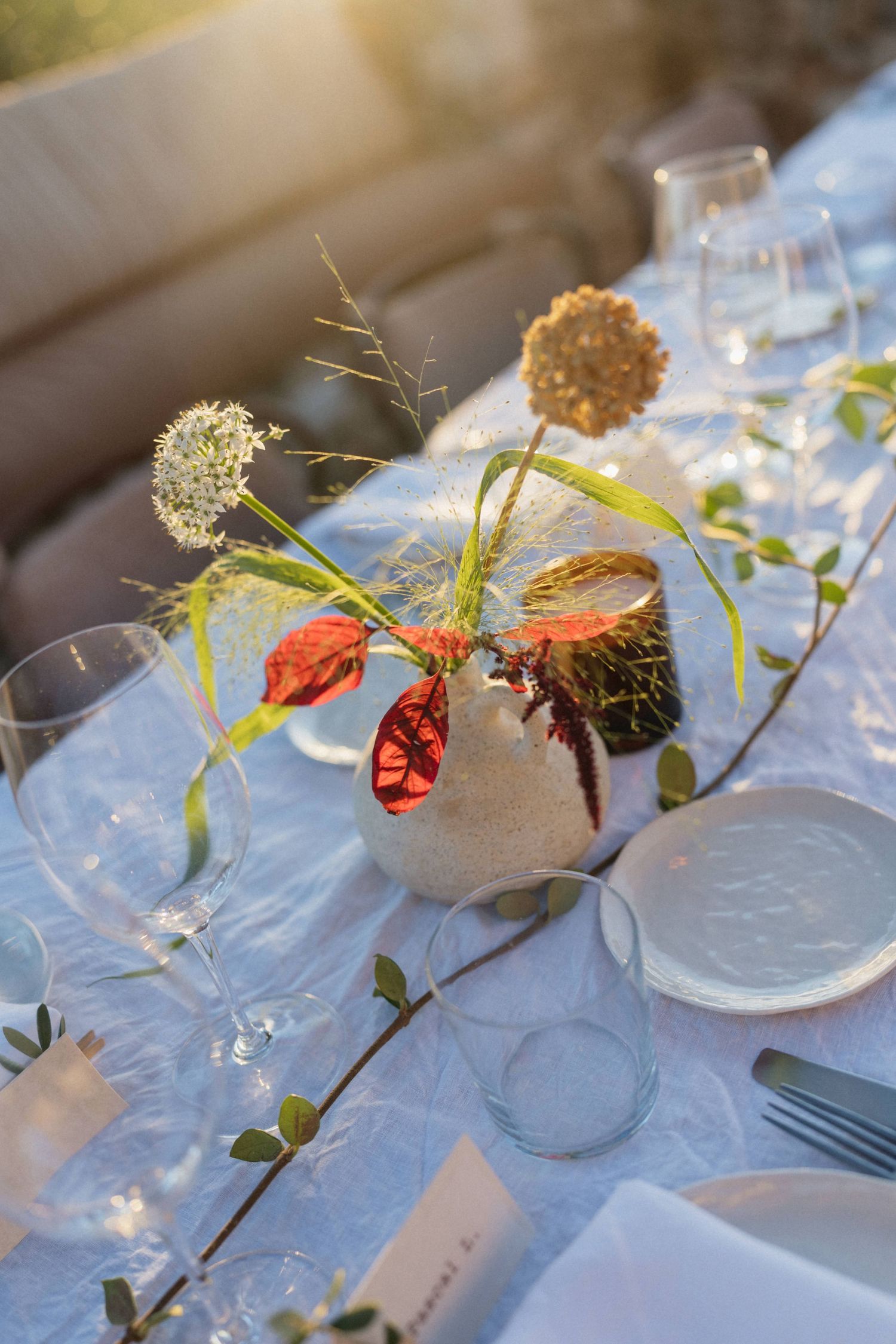 Wedding table centerpiece with dried flowers and red leaves in a small white vase beside glassware and place cards.