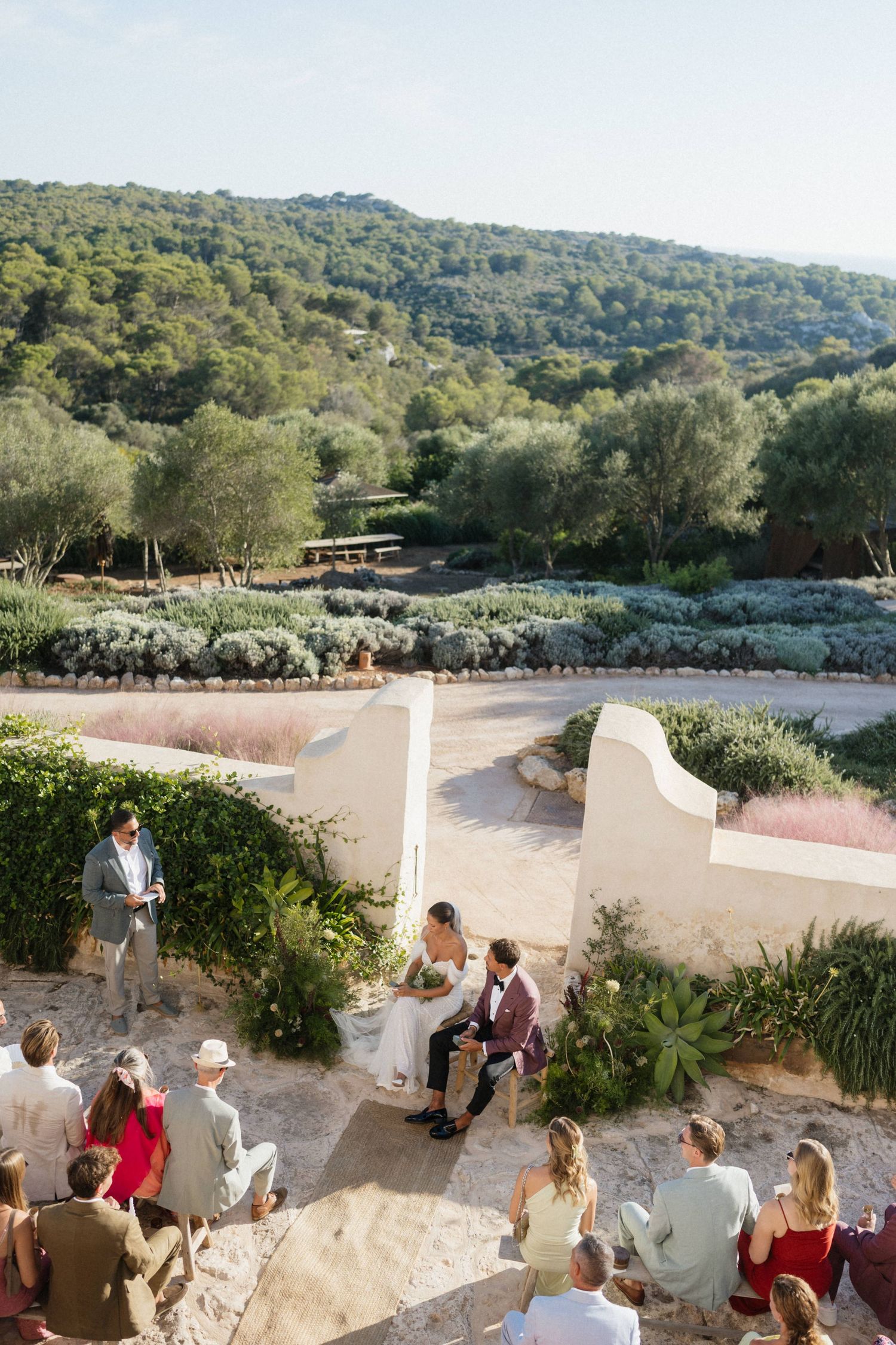 Outdoor wedding ceremony with guests seated on stone steps as the couple sits facing the officiant among Mediterranean greenery.