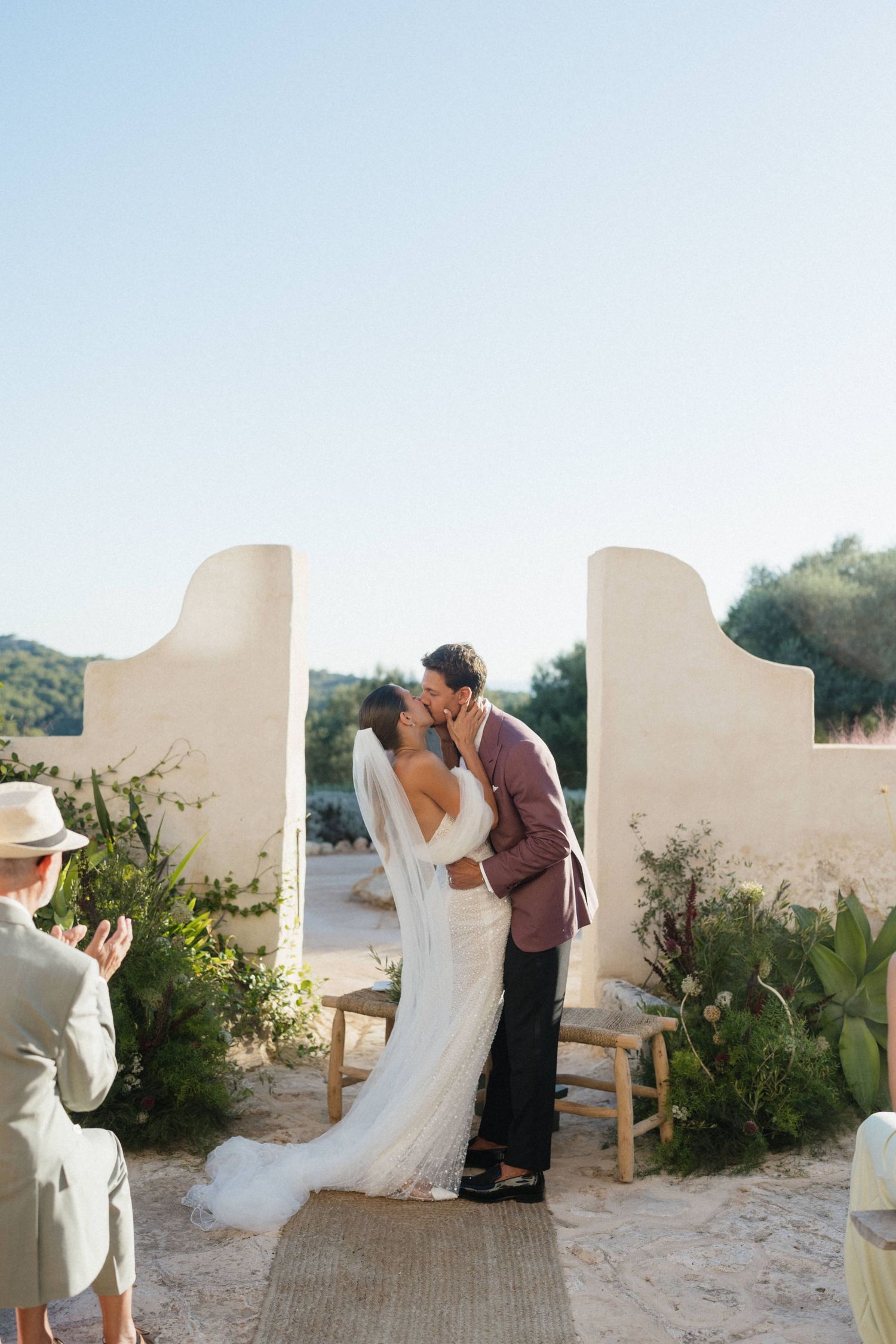 Bride in a flowing white gown and veil kisses her groom in a burgundy blazer during an outdoor ceremony between white stucco walls.