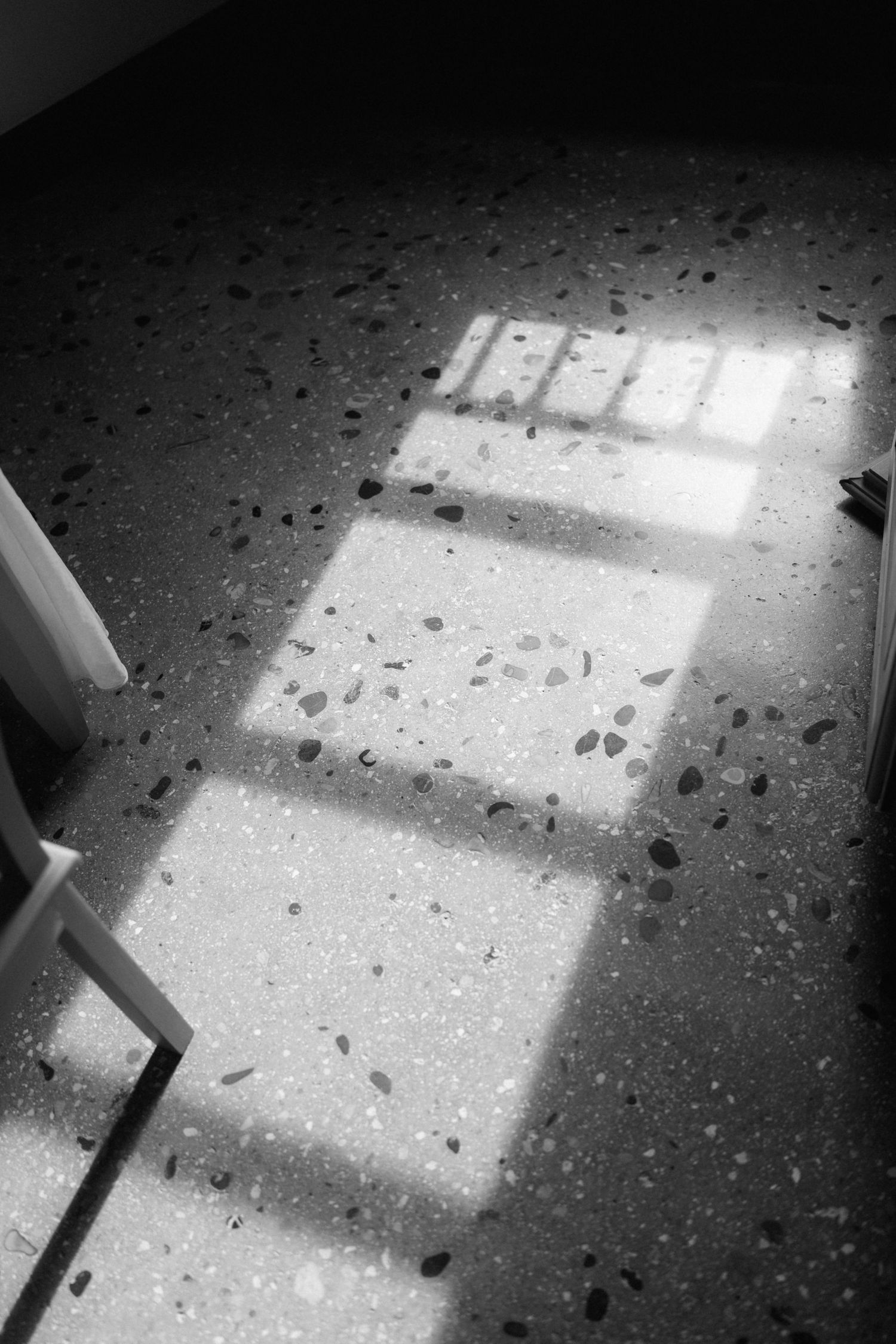 Black and white photo of sunlight casting window shadows across a speckled terrazzo floor beside a white chair.