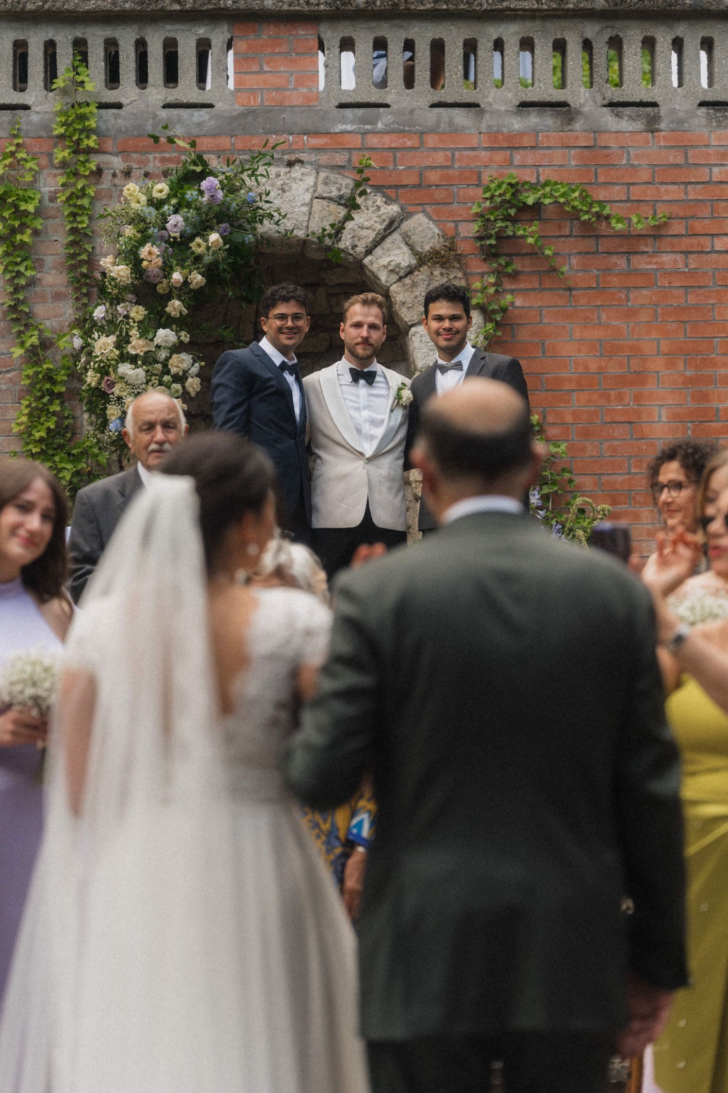 Groom awaiting bride at floral arch altar during outdoor wedding ceremony at brick venue.