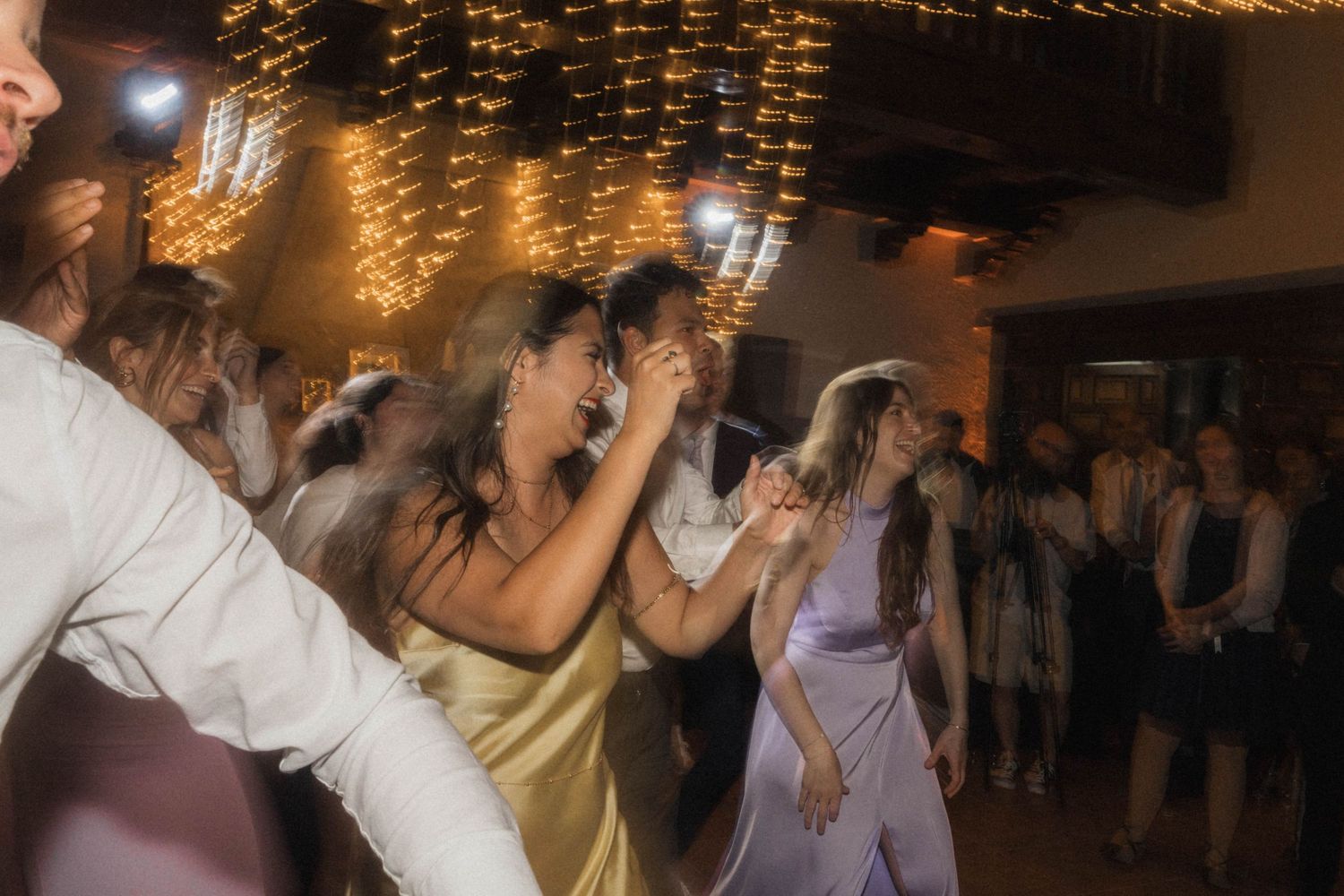 Energetic wedding guests dancing under warm string lights at lively reception celebration.