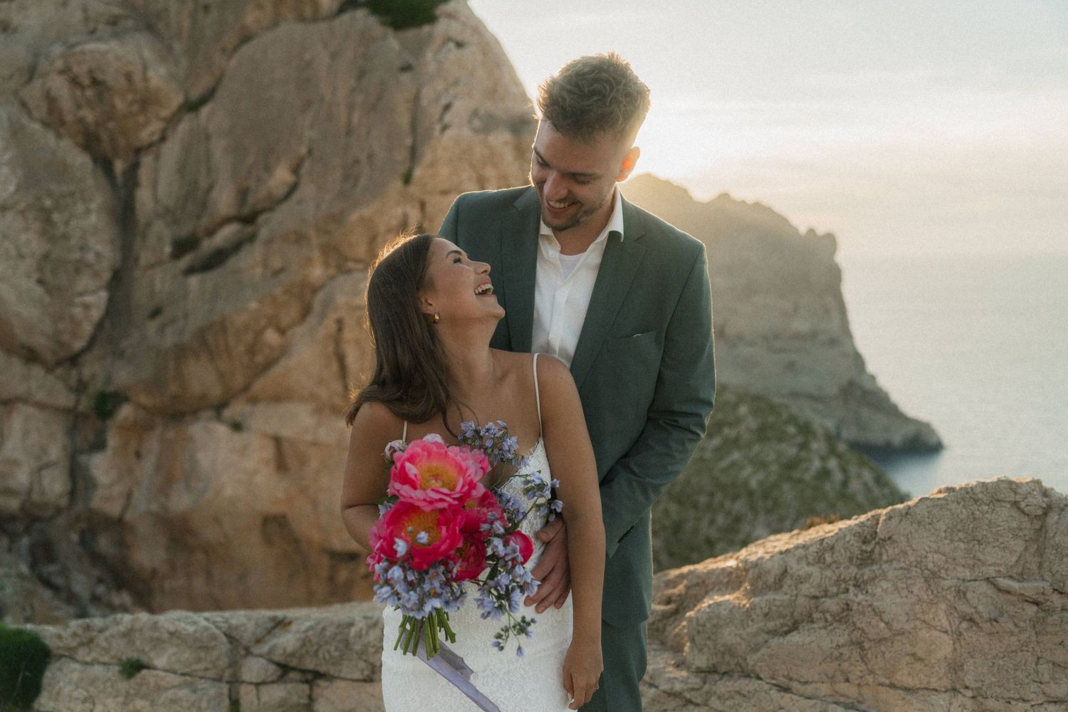 Bride and groom share a romantic moment on rocky cliffs at sunset, bride holding vibrant pink peony bouquet.