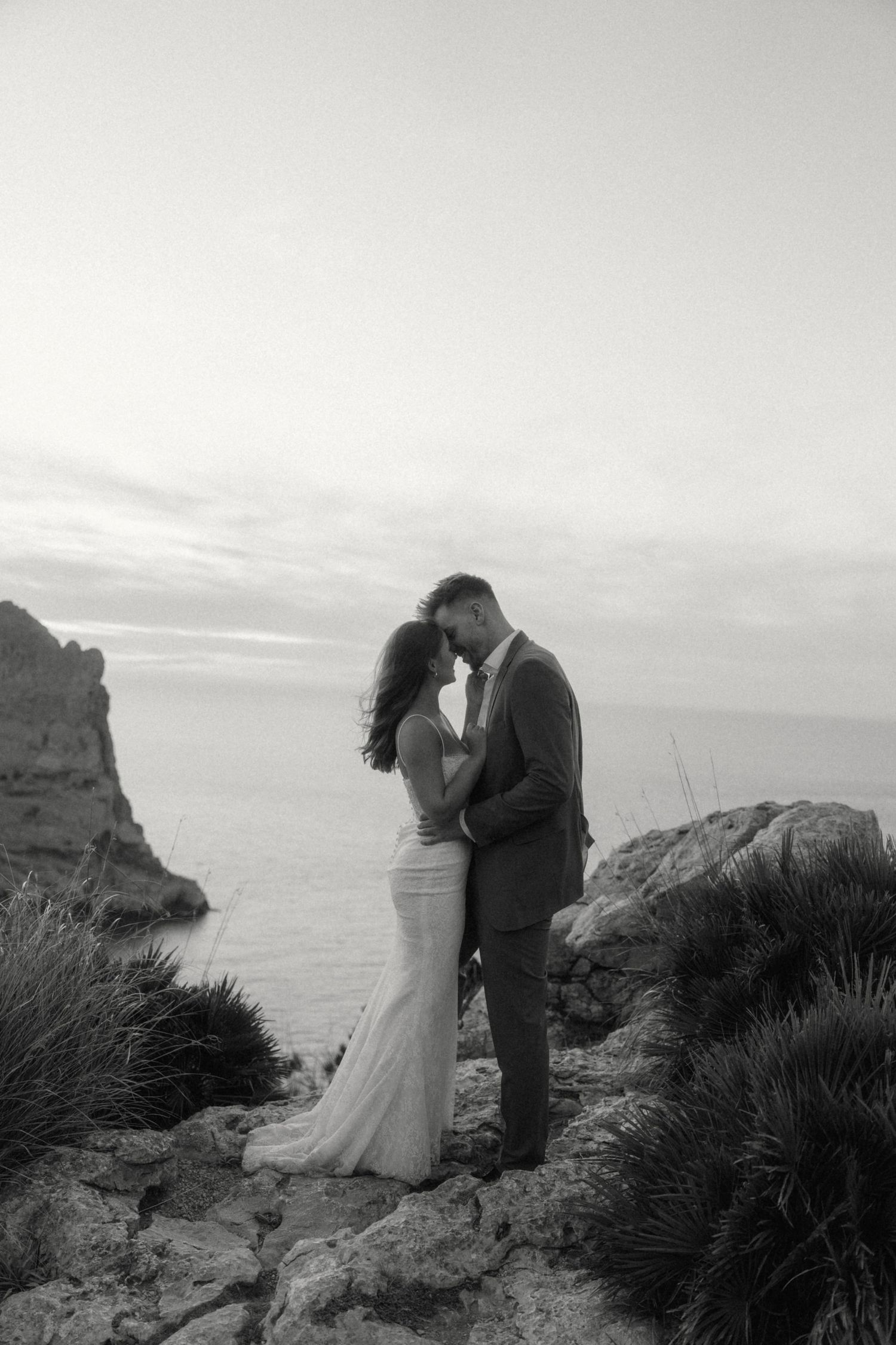 Black and white photo of bride and groom embracing on rocky seaside cliffs at dusk.