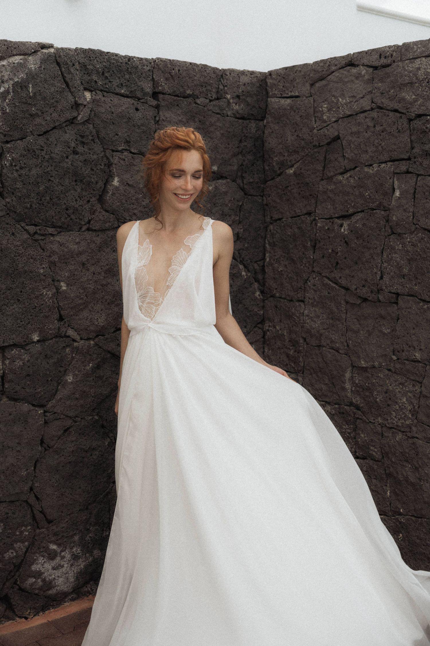Bride in deep V-neck white flowing wedding gown standing against dark volcanic stone wall.