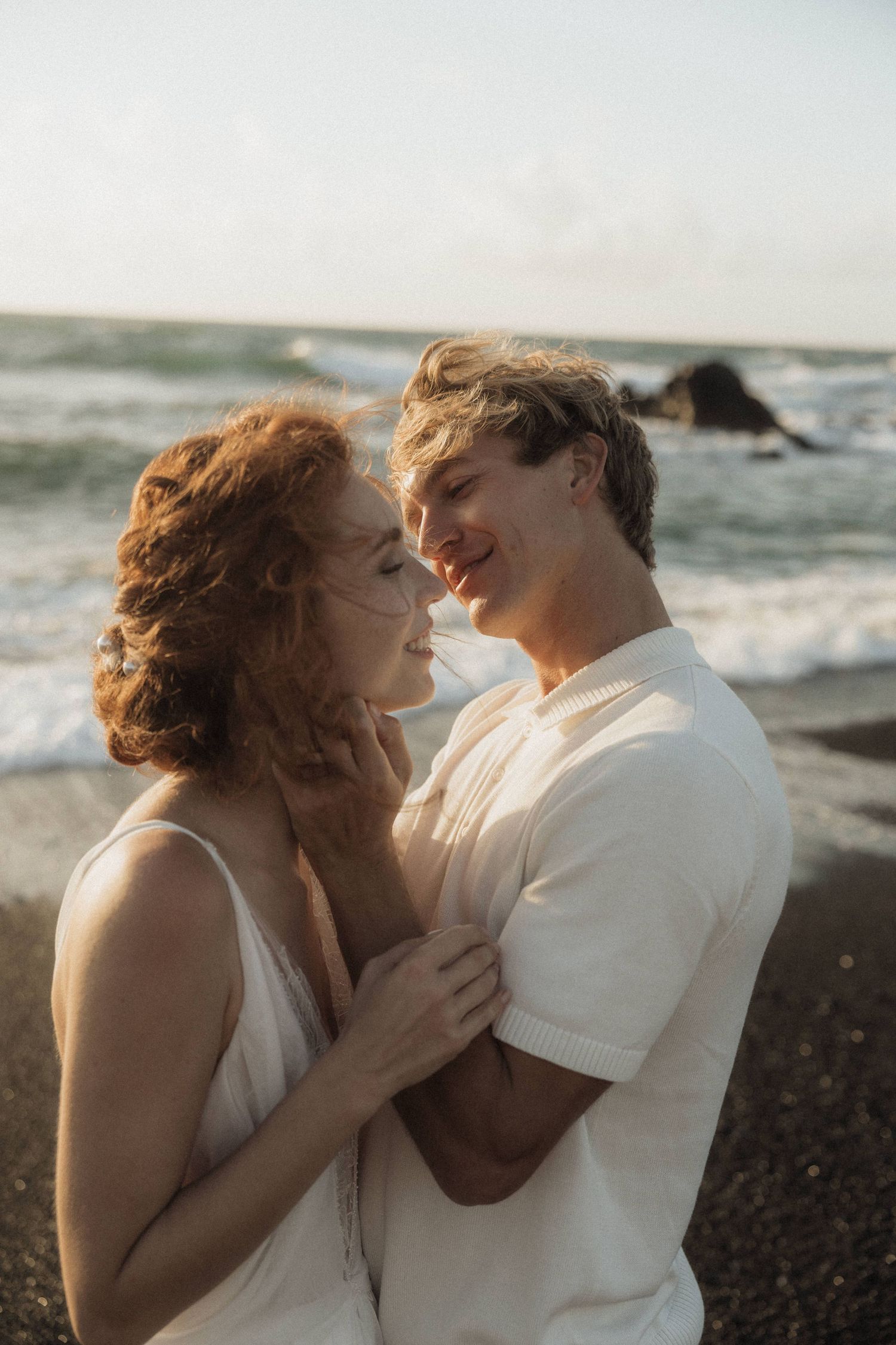 Romantic redhead bride and groom embracing and smiling on black sand beach at sunset.