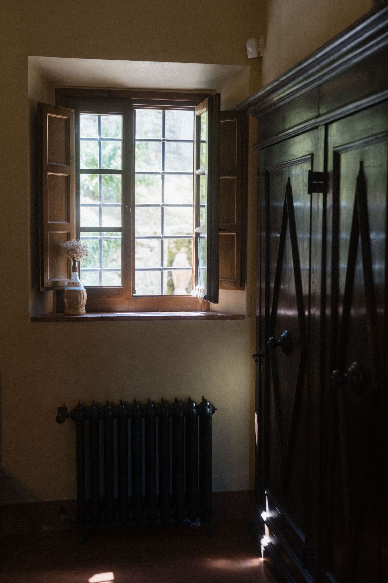Rustic interior room with an open wooden window, antique radiator, and dark wood armoire in warm light.