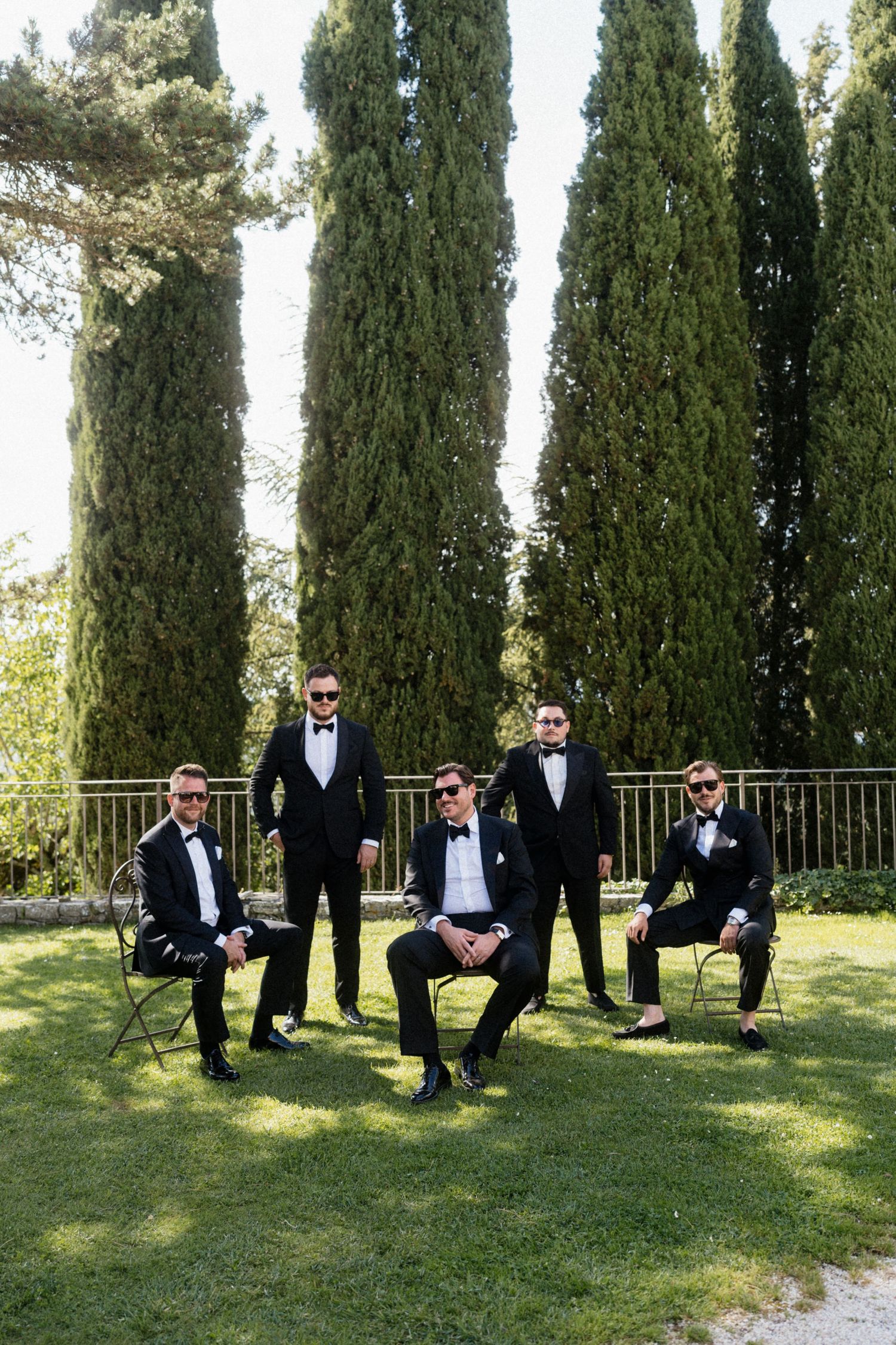 Five groomsmen in black tuxedos and bow ties posing with cypress trees in a formal Italian garden.