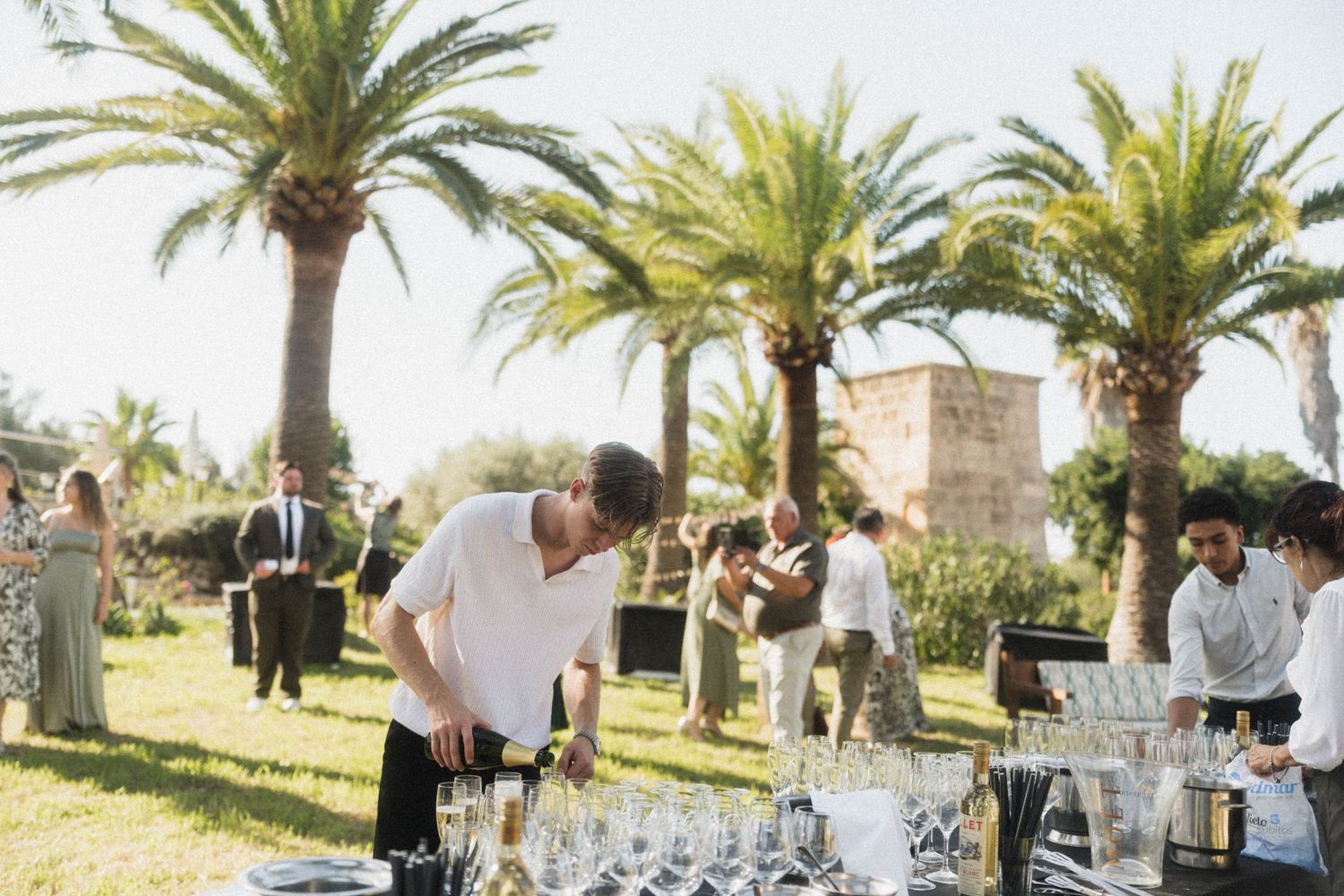 Wedding staff preparing champagne glasses at outdoor reception under palm trees.