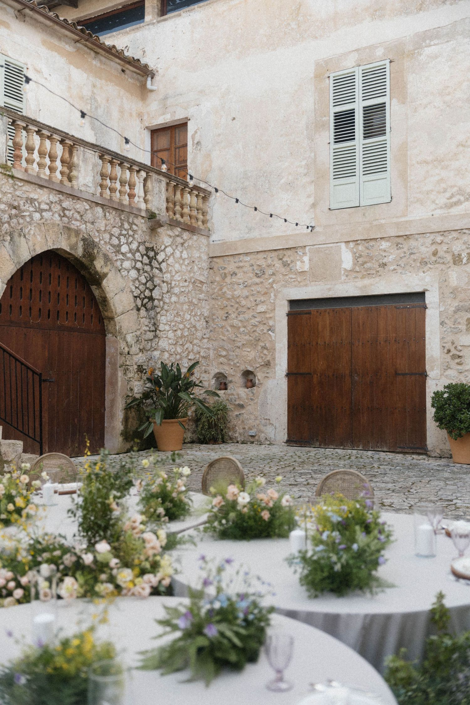 Romantic outdoor wedding reception with floral centerpieces in a historic stone courtyard with wooden arched doors.