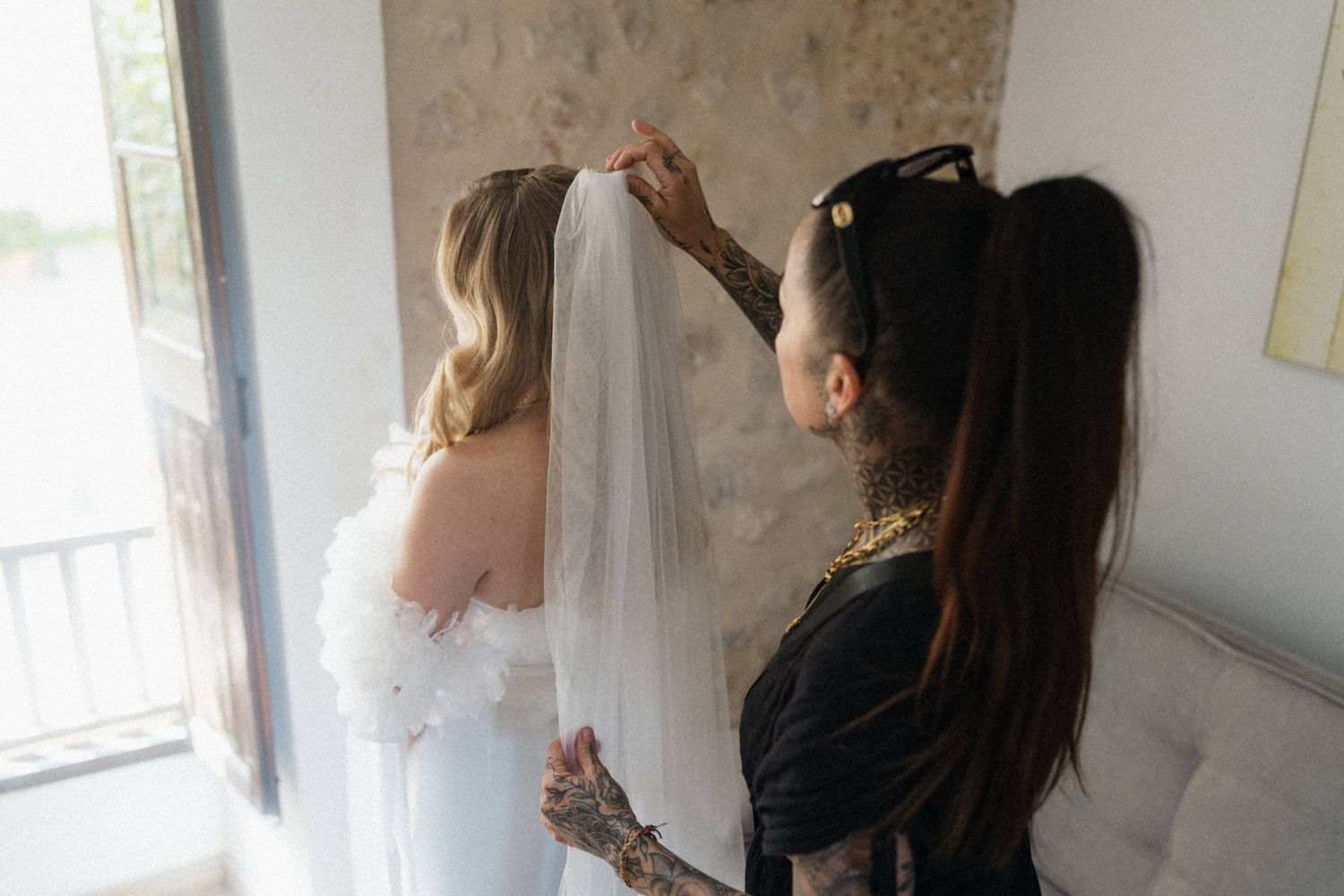 Tattooed bridesmaid adjusting bridal veil for blonde bride in white wedding dress near window.