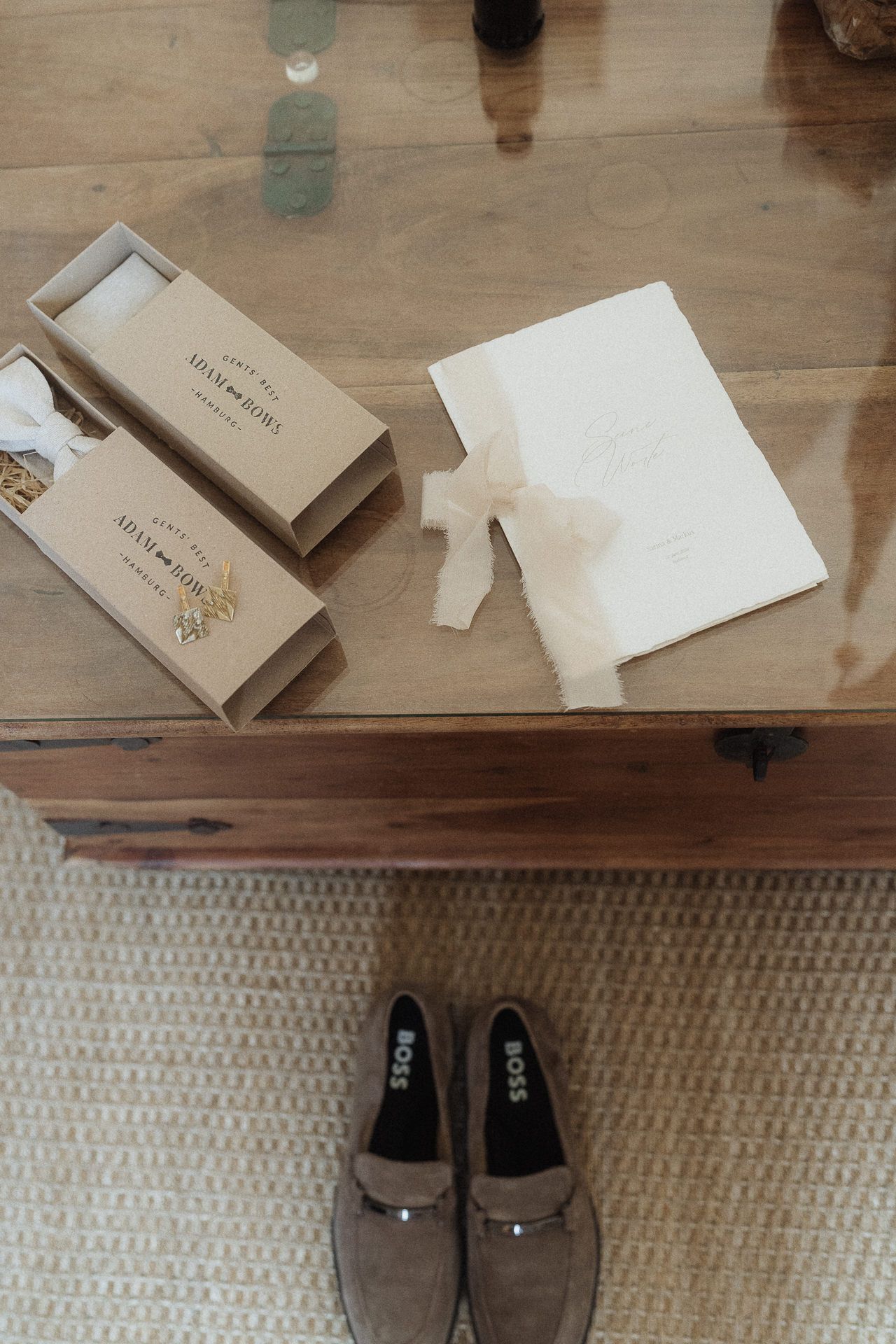 Overhead view of wedding stationery with velvet ribbon, kraft gift boxes, and BOSS loafers on a wooden table.