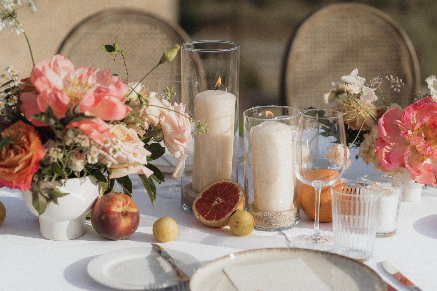 Elegant wedding table setting with pink peonies, pillar candles, citrus fruits, and rose gold glassware on white linen.