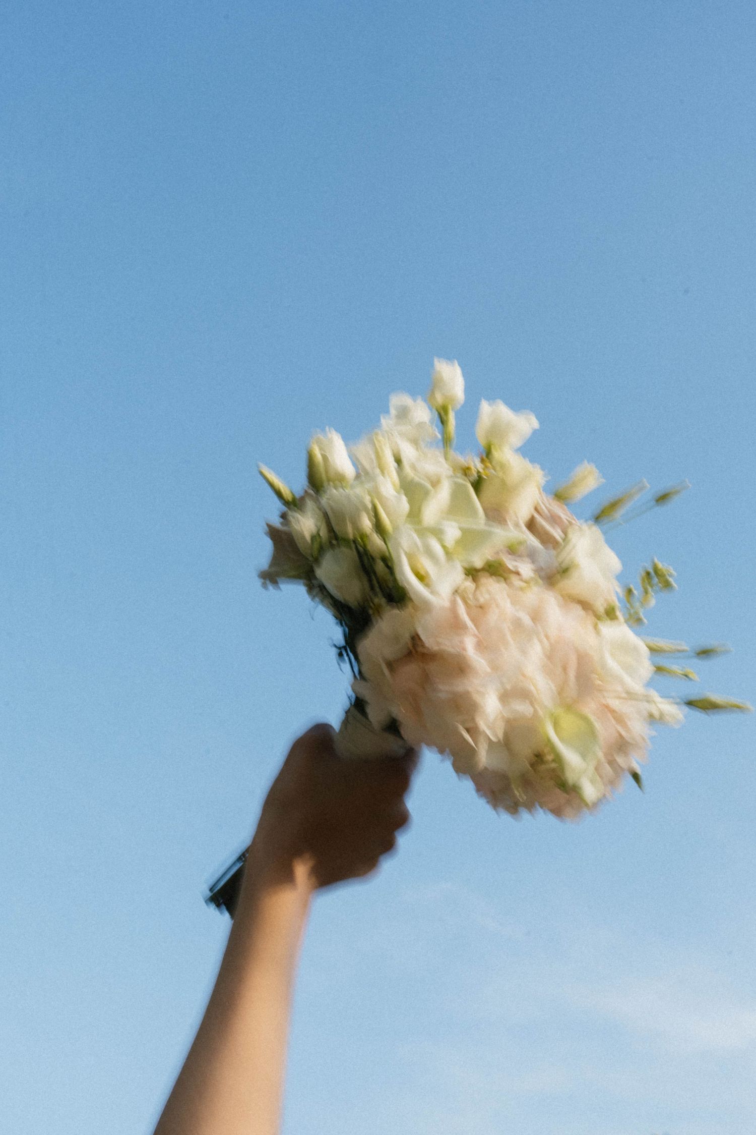 A hand holds a white bridal bouquet of roses and eustoma flowers raised against a clear blue sky.