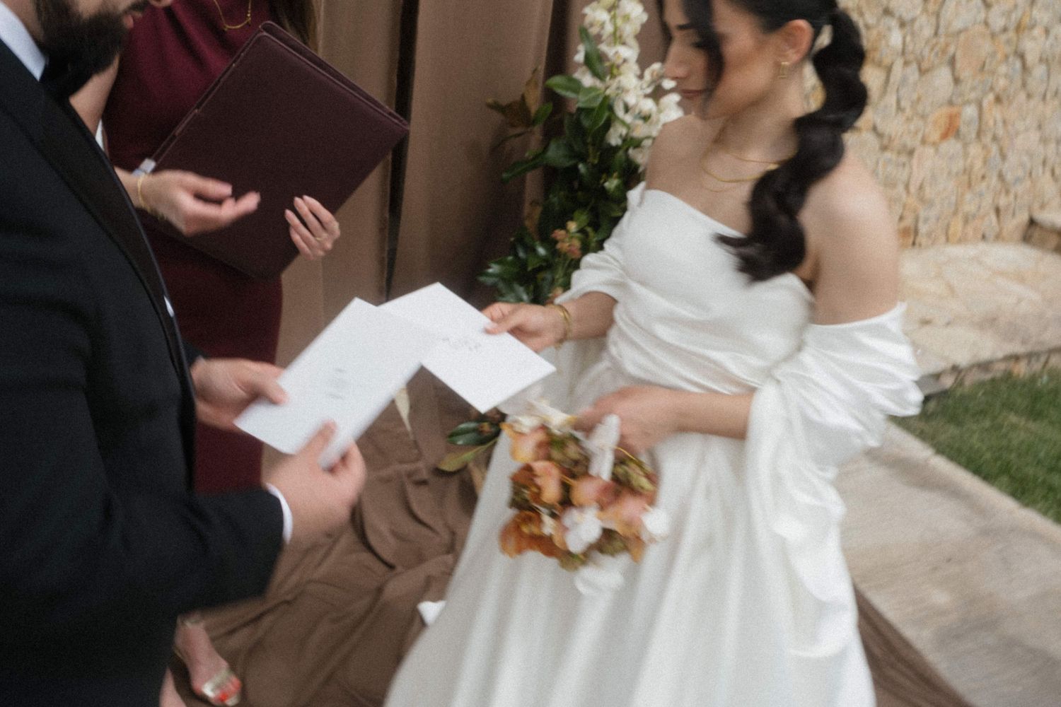Bride in off-shoulder white gown holding colorful bouquet reading vows during outdoor wedding ceremony.