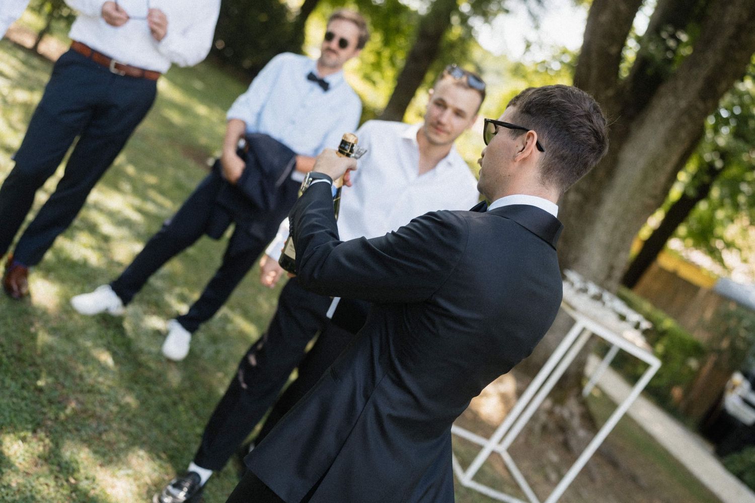 Groom in black suit toasting with guests at an outdoor wedding reception in a green park setting.
