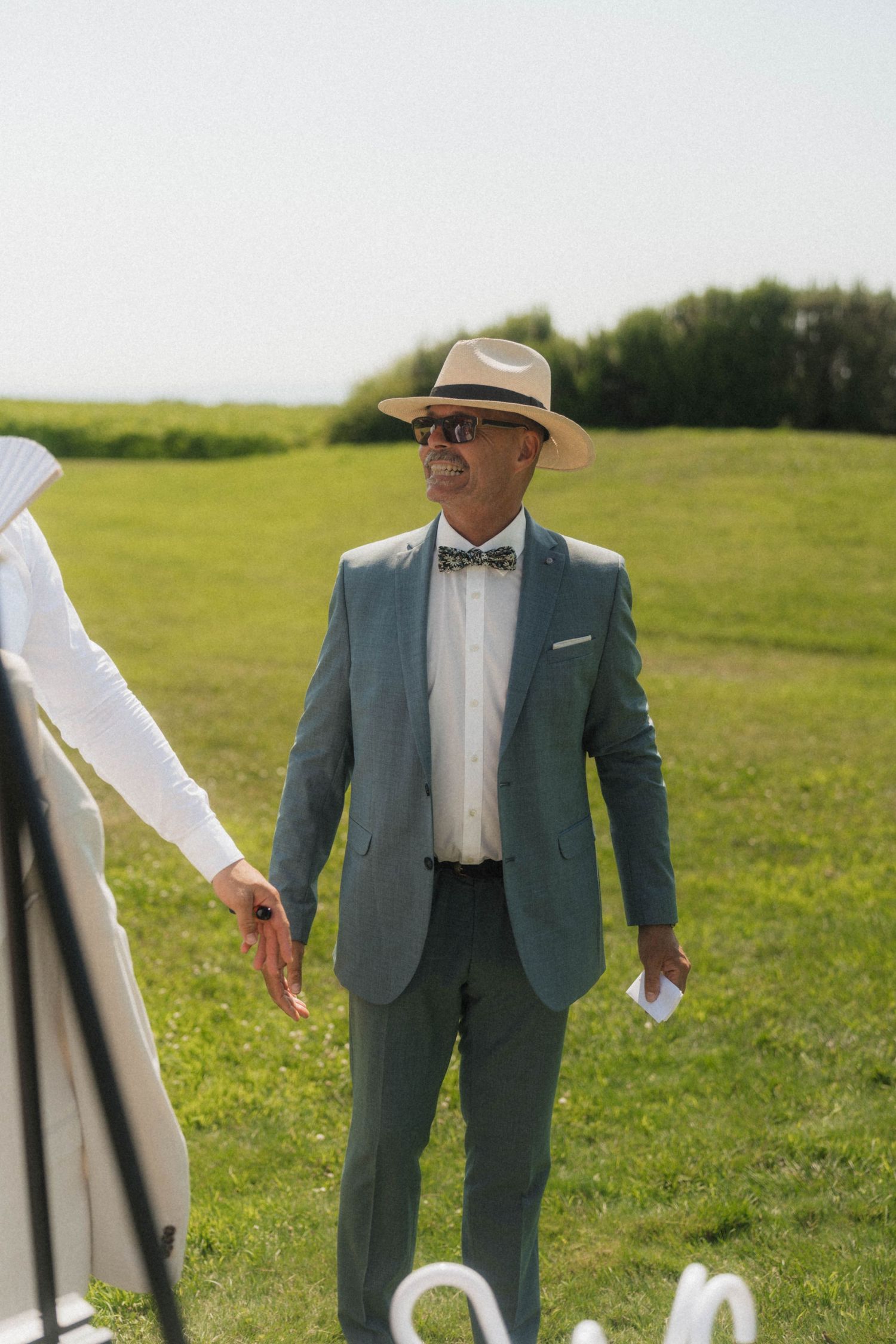 Stylish man in gray suit, bow tie, and panama hat standing on green lawn at outdoor wedding.