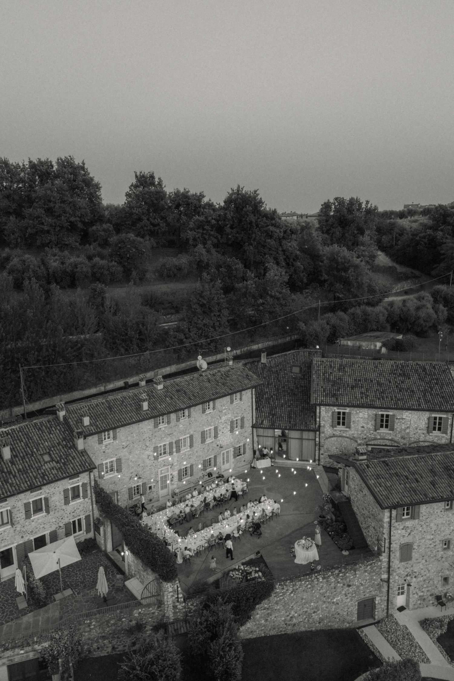 Black and white aerial view of illuminated outdoor wedding reception at rustic Italian villa.