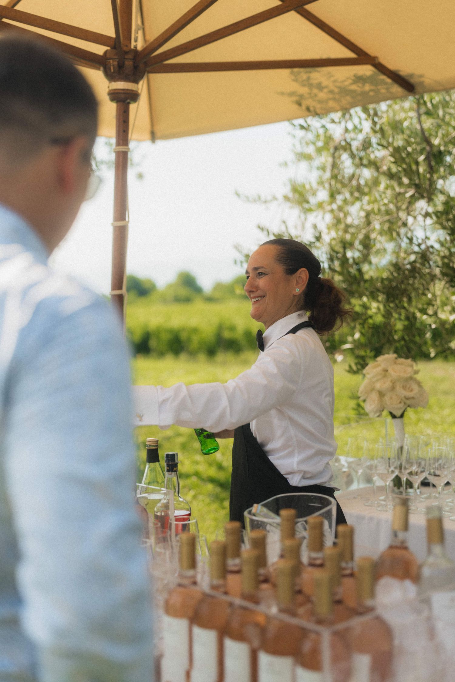 Smiling female bartender serves drinks at an outdoor wedding bar surrounded by wine bottles and glassware.