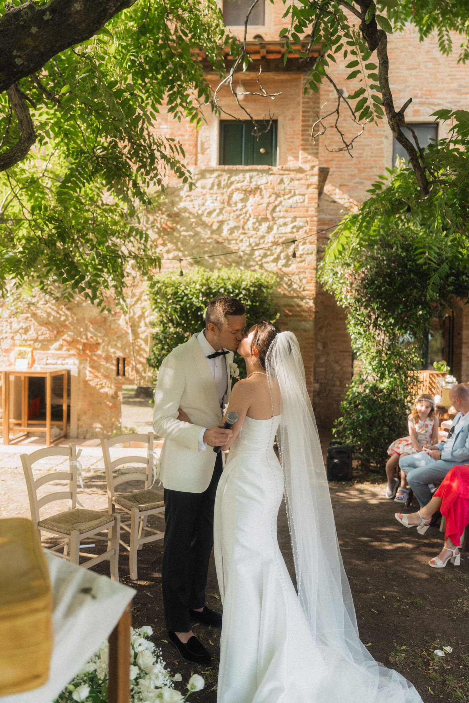 Bride and groom share first kiss at outdoor wedding ceremony beside rustic stone building in Tuscany.