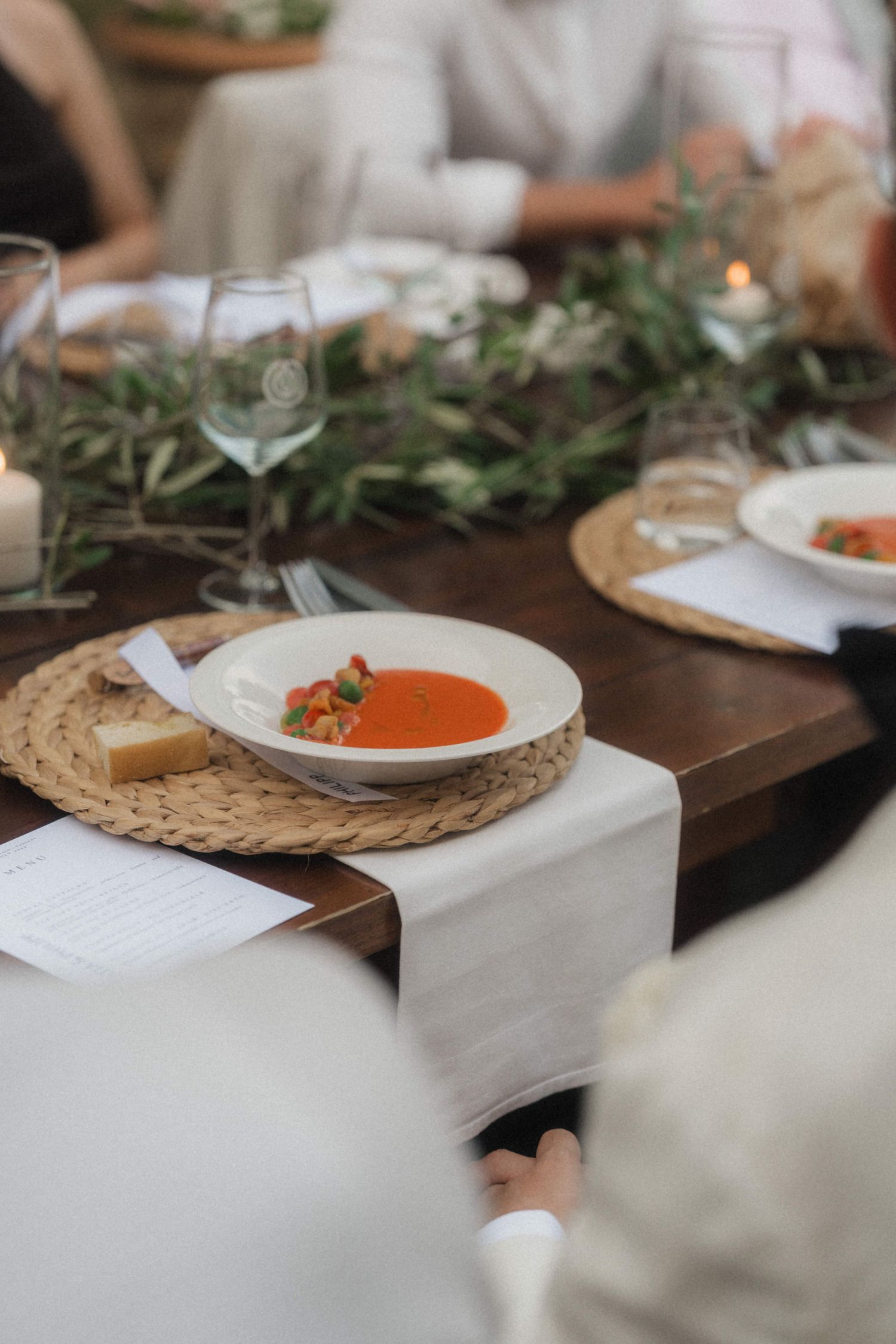Elegant wedding table setting with tomato soup, woven placemats, glassware, and greenery centerpiece.