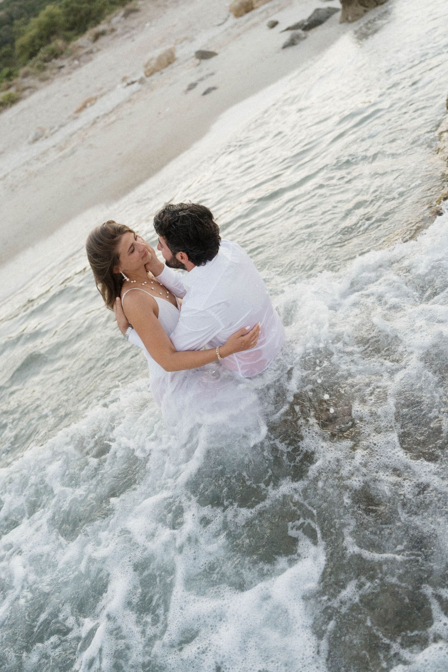 Wedding couple embracing in crashing ocean waves, wearing white outfits, captured from above on beach.
