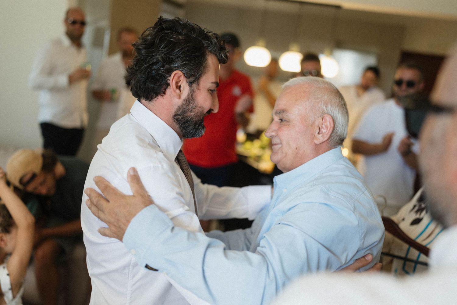 Two men share a warm embrace at an indoor social gathering, one in a white shirt with a beard.