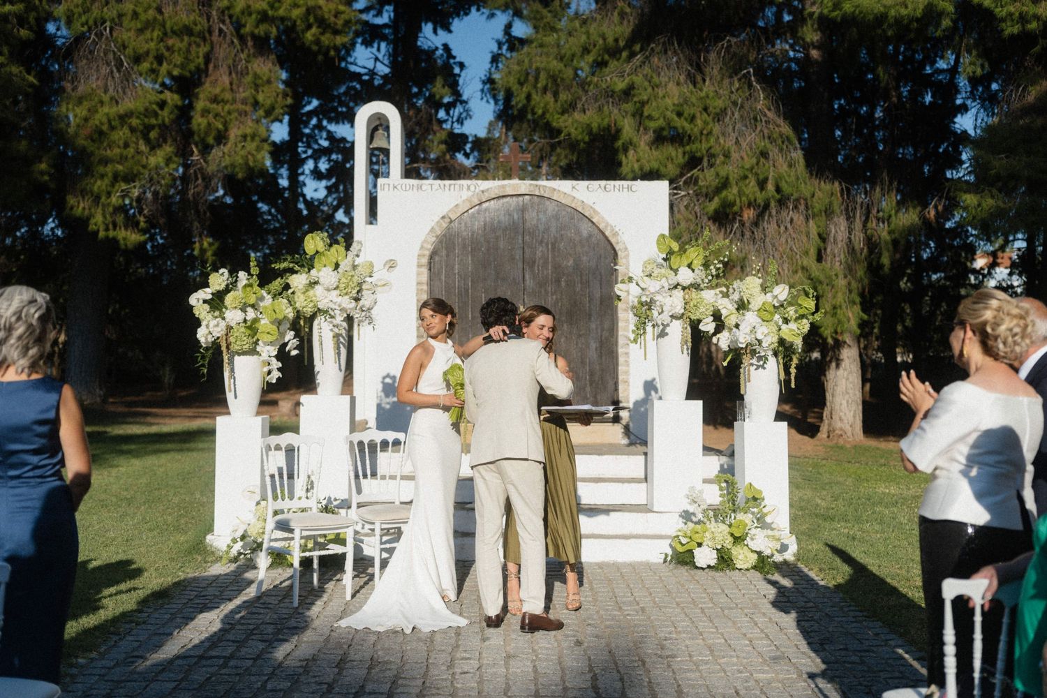 Couple exchanges vows at elegant outdoor wedding ceremony with white floral arch and chapel backdrop.