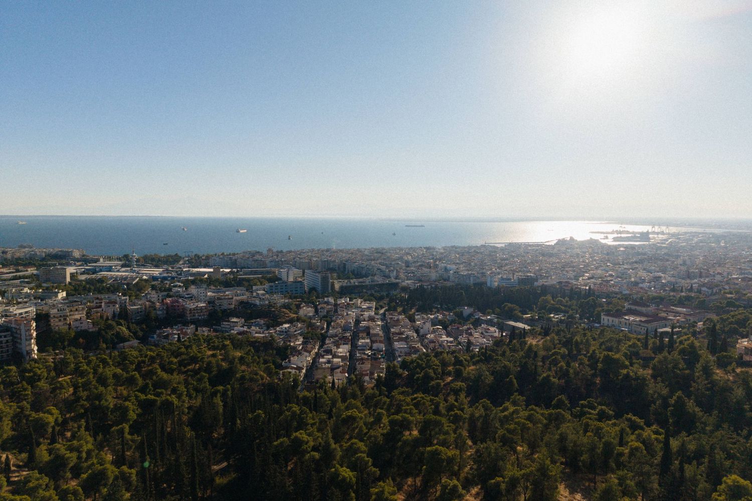 Aerial panoramic view of Thessaloniki city with dense urban landscape, lush green trees, and the blue Aegean Sea in the background.