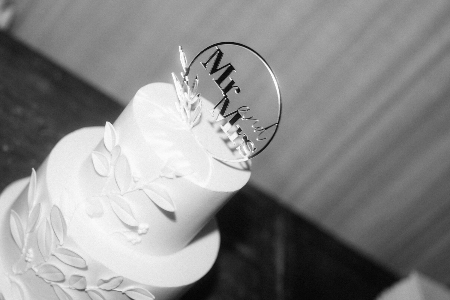 Black and white close-up of a tiered white wedding cake topped with an elegant Mr and Mrs acrylic cake topper.