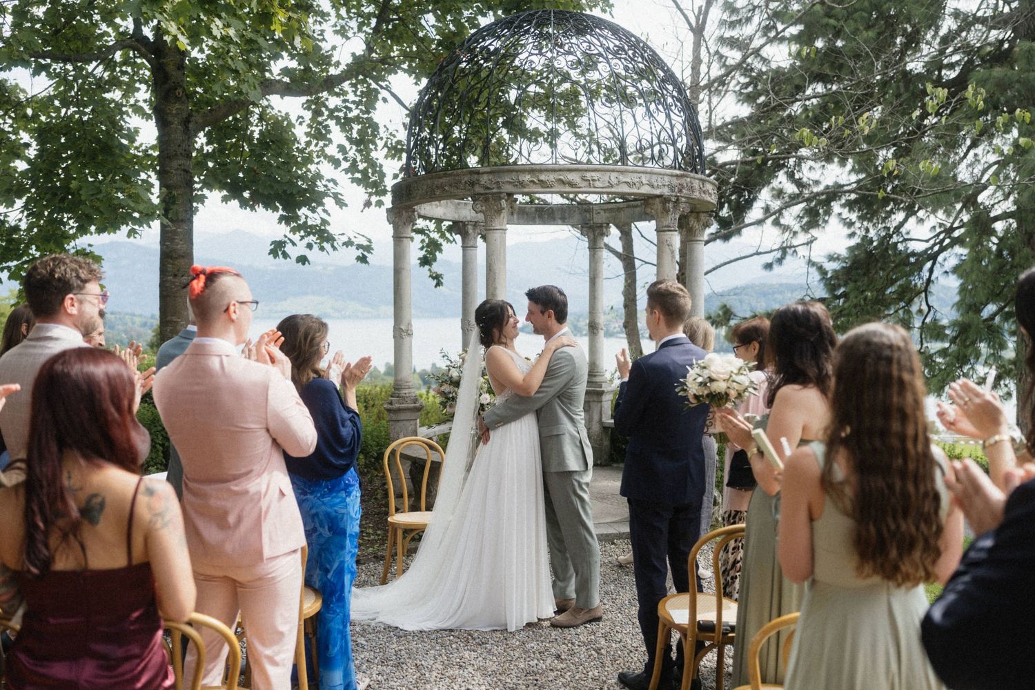 Newlyweds share first kiss under ornate stone gazebo as wedding guests applaud in celebration.