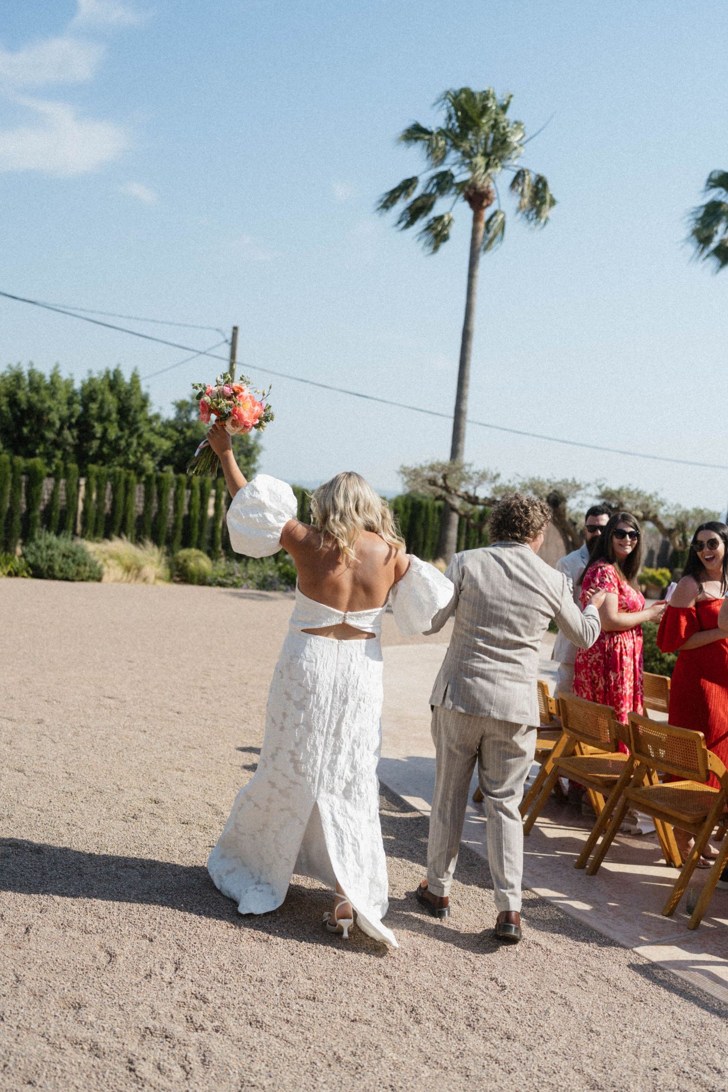 Bride in white dress raises bouquet while walking with groom on sandy outdoor wedding venue with palm trees.